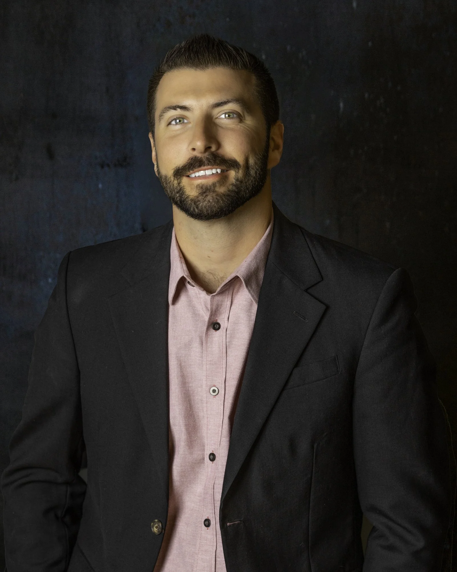 Man with dark hair and beard wearing a black suit jacket and pink shirt, smiling against a dark background.