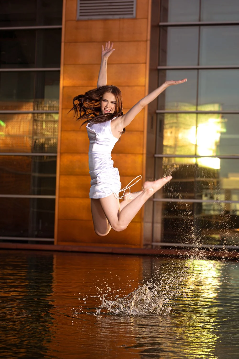 A woman in a white dress jumping joyfully over water, with a splash below her, in front of a modern building with glass windows.