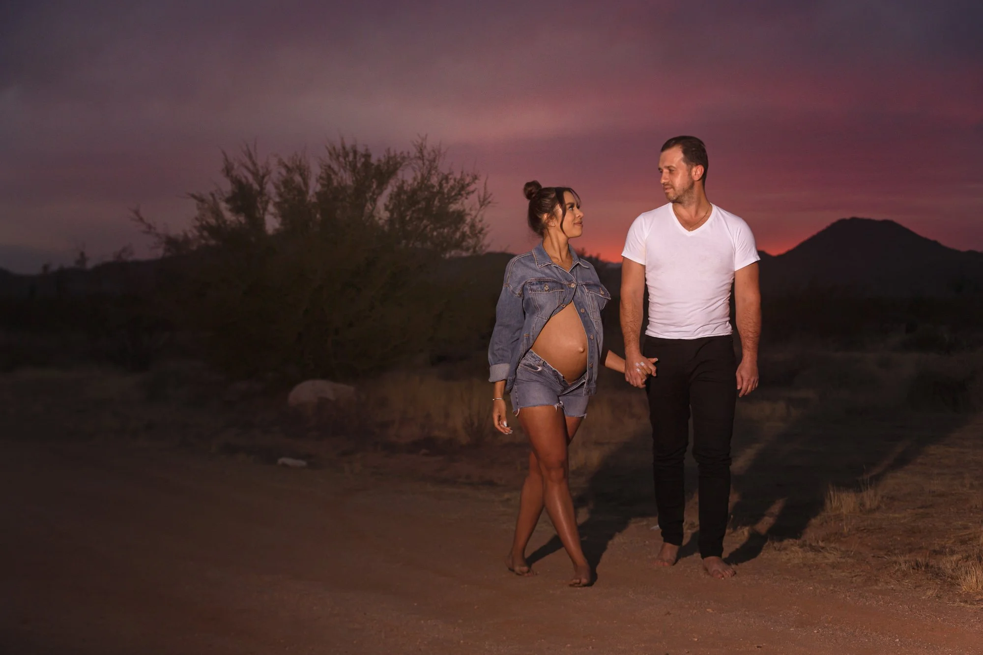 A pregnant woman and a man holding hands, walking on a dirt path with a sunset backdrop. The woman is wearing a denim jacket and shorts, and the man is in a white t-shirt and black pants. Desert landscape surrounds them.