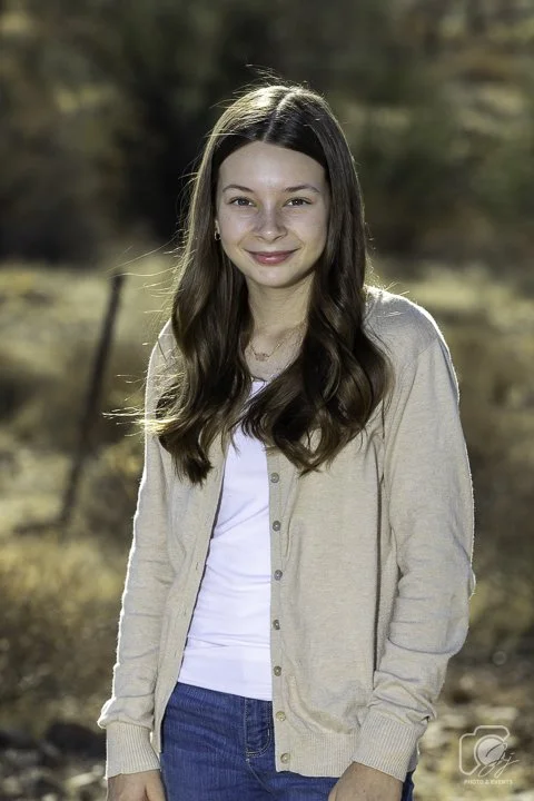 Young woman wearing a beige sweater and jeans, standing outdoors with blurred natural background.