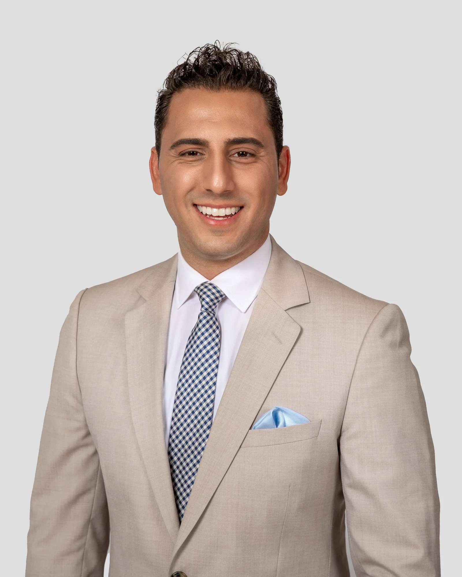 Smiling man in a light brown suit with a blue checkered tie and a light blue pocket square, against a gray background.