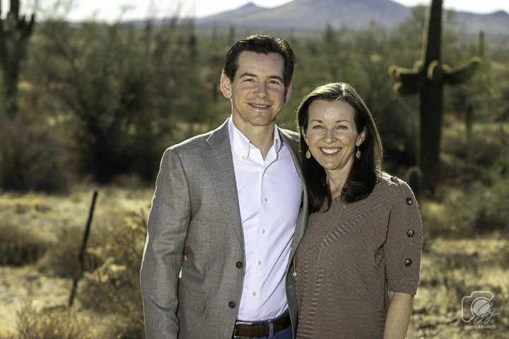 A man and a woman standing together in a desert landscape, with cacti and mountains in the background. The man is wearing a gray suit jacket and white shirt, while the woman is wearing a brown dress with button details and long earrings. They are both smiling at the camera.