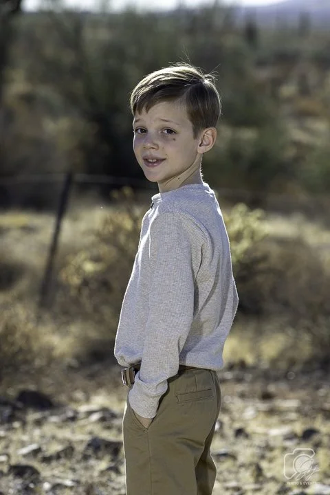 Young boy in a desert setting, wearing a light gray long-sleeve shirt and khaki pants, standing with hands in pockets.