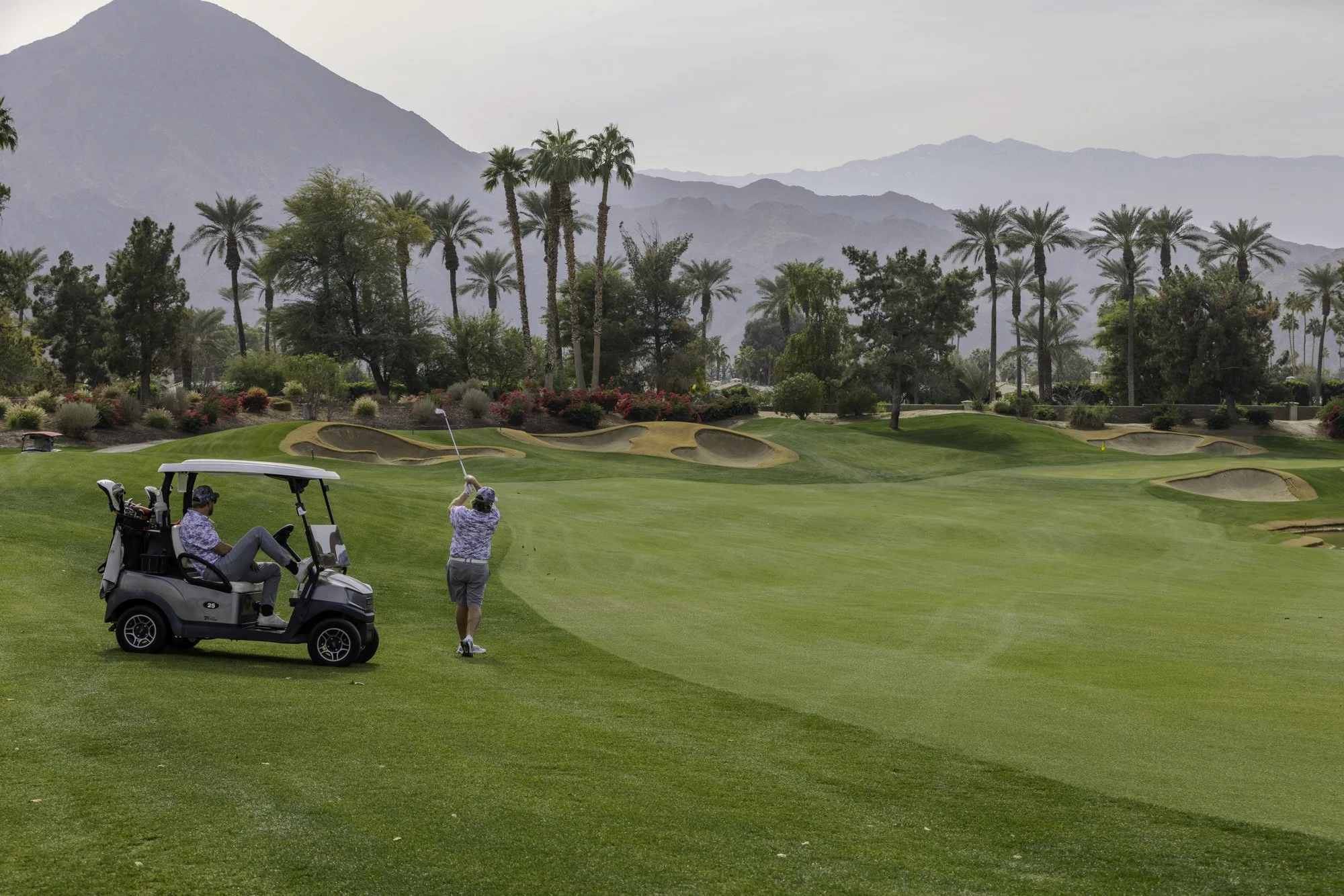 Golfer swinging club on a lush golf course with palm trees and mountains in the background, alongside a parked golf cart.