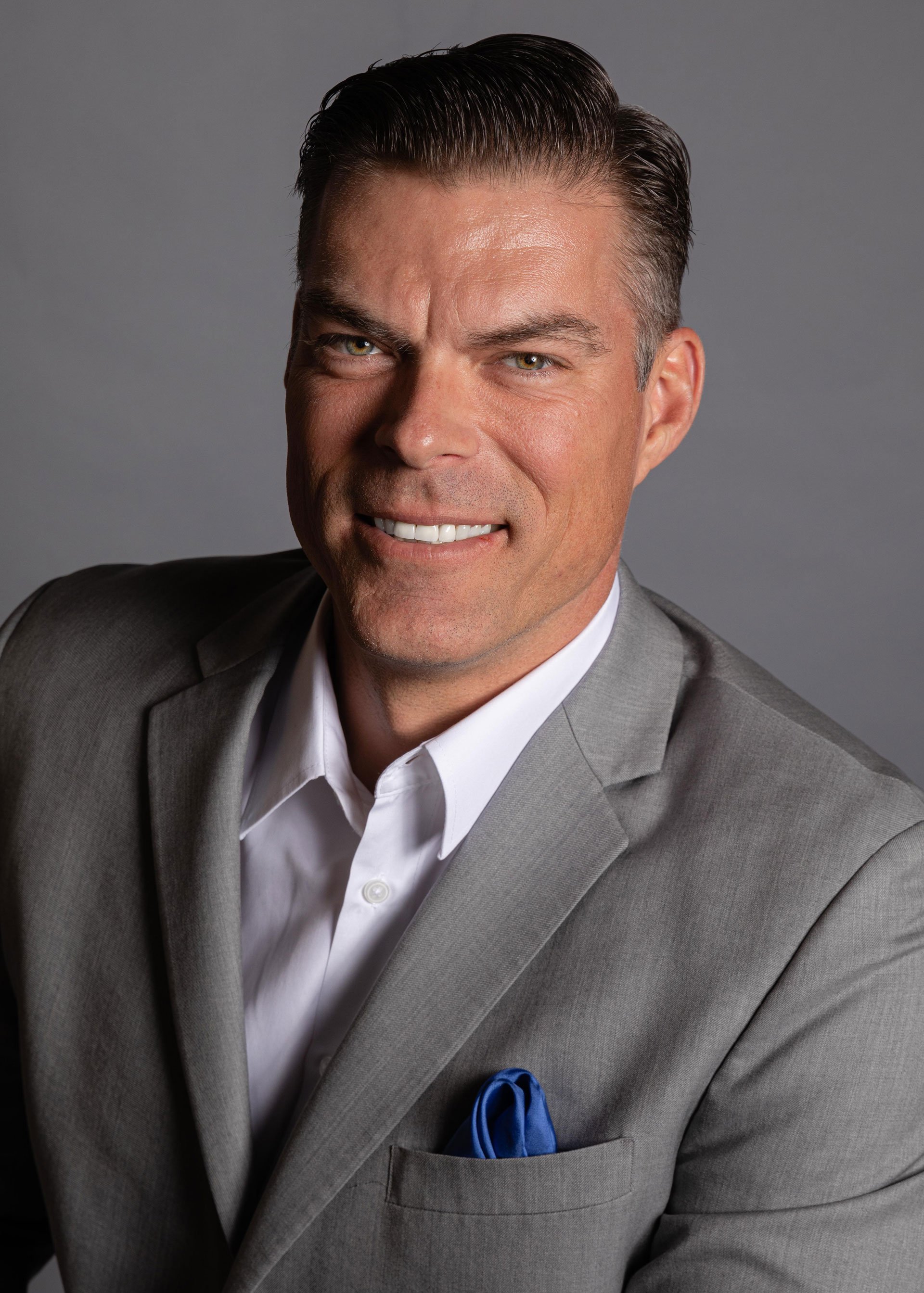 Man in a gray suit with white shirt and blue pocket square, smiling.