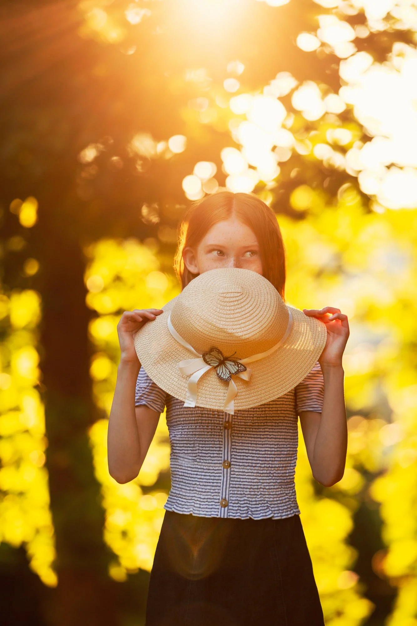 Girl holding a straw hat with a butterfly decoration, standing in a sunlit forest.