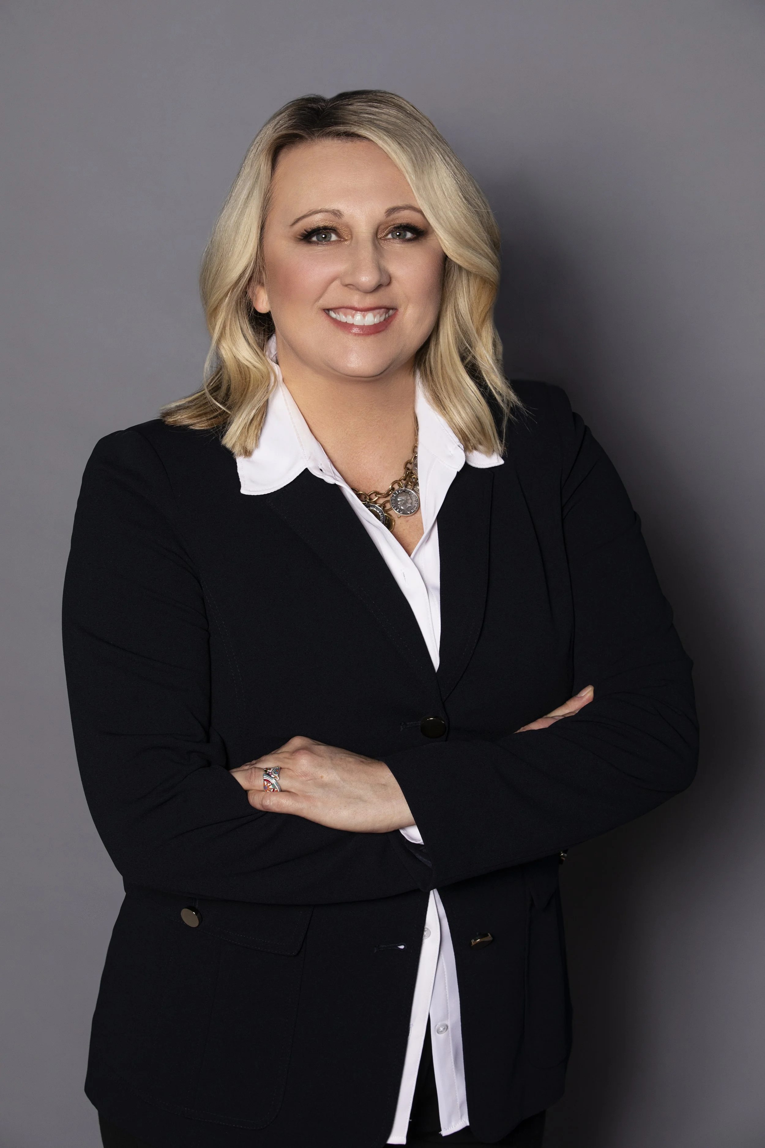 Woman in business attire with arms crossed, smiling against gray background.