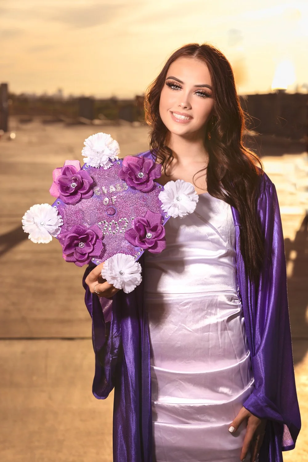 Young woman in graduation attire holding a decorated cap with "Class of 2023" and flowers