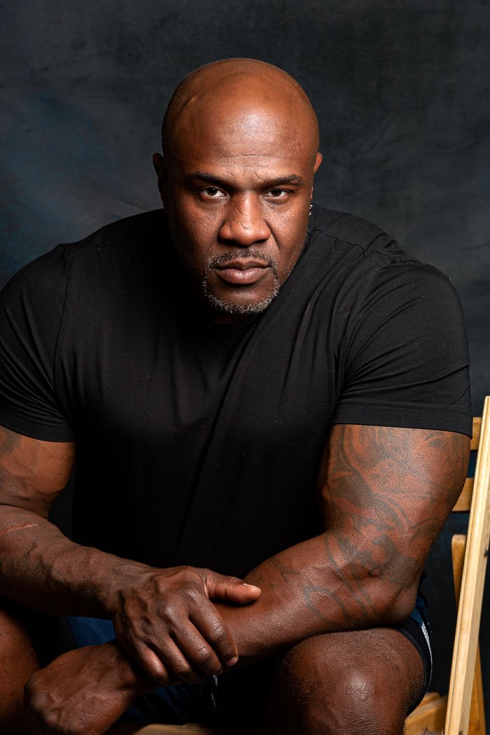 Man in black t-shirt with tattoos sitting on a chair with a serious expression against a dark background.