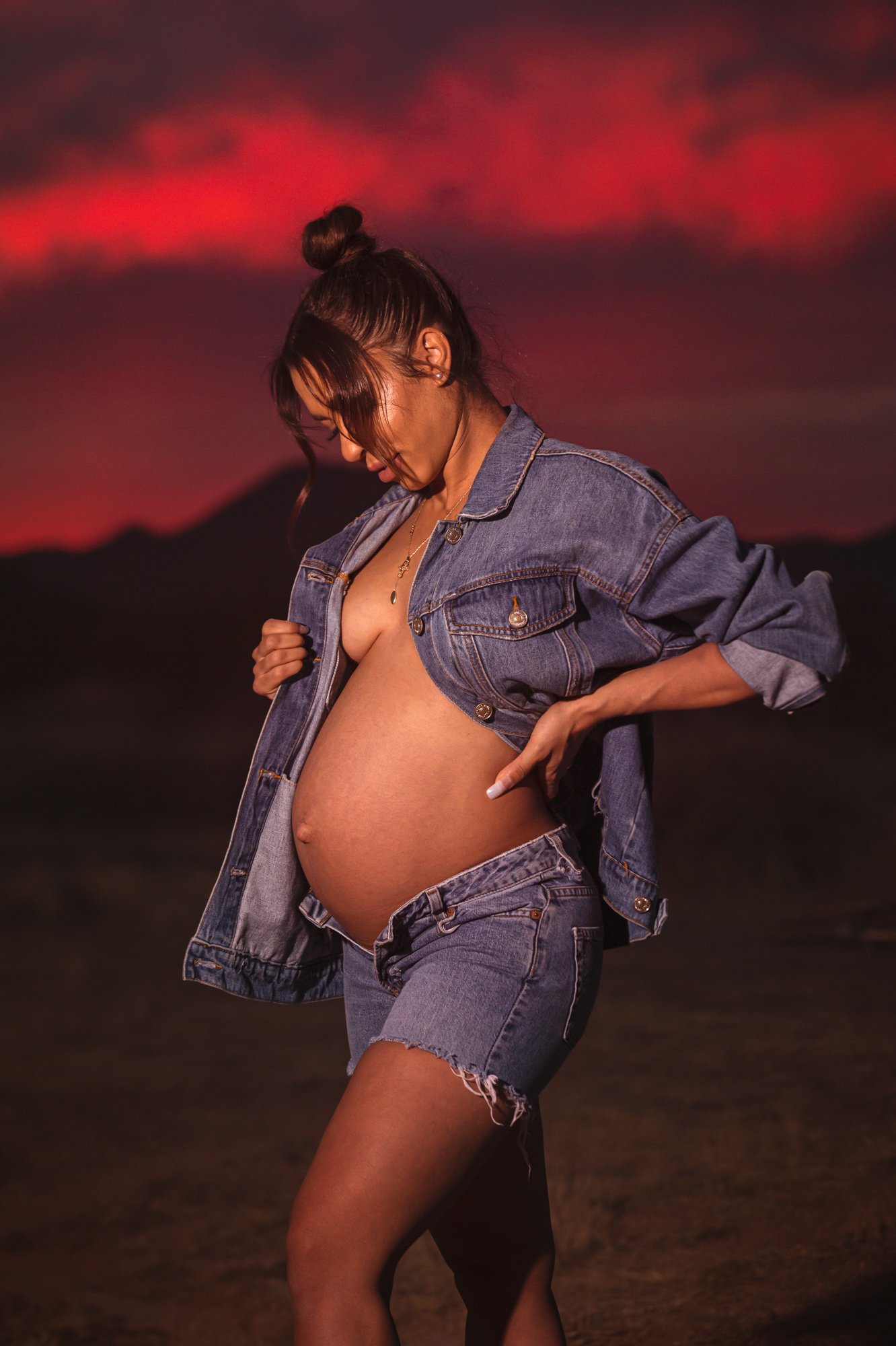 Pregnant woman in denim jacket and shorts at sunset.