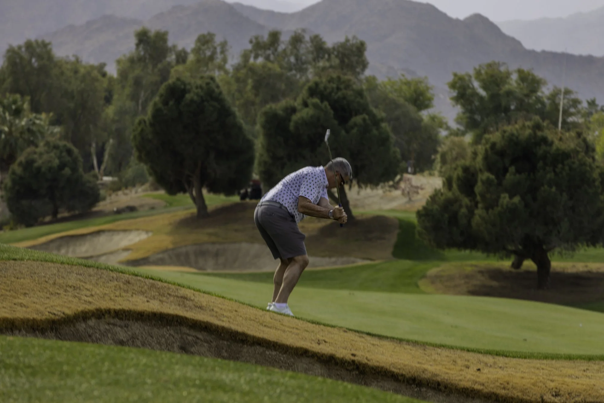 Golfer swinging club on grassy hill with trees and mountains in background.