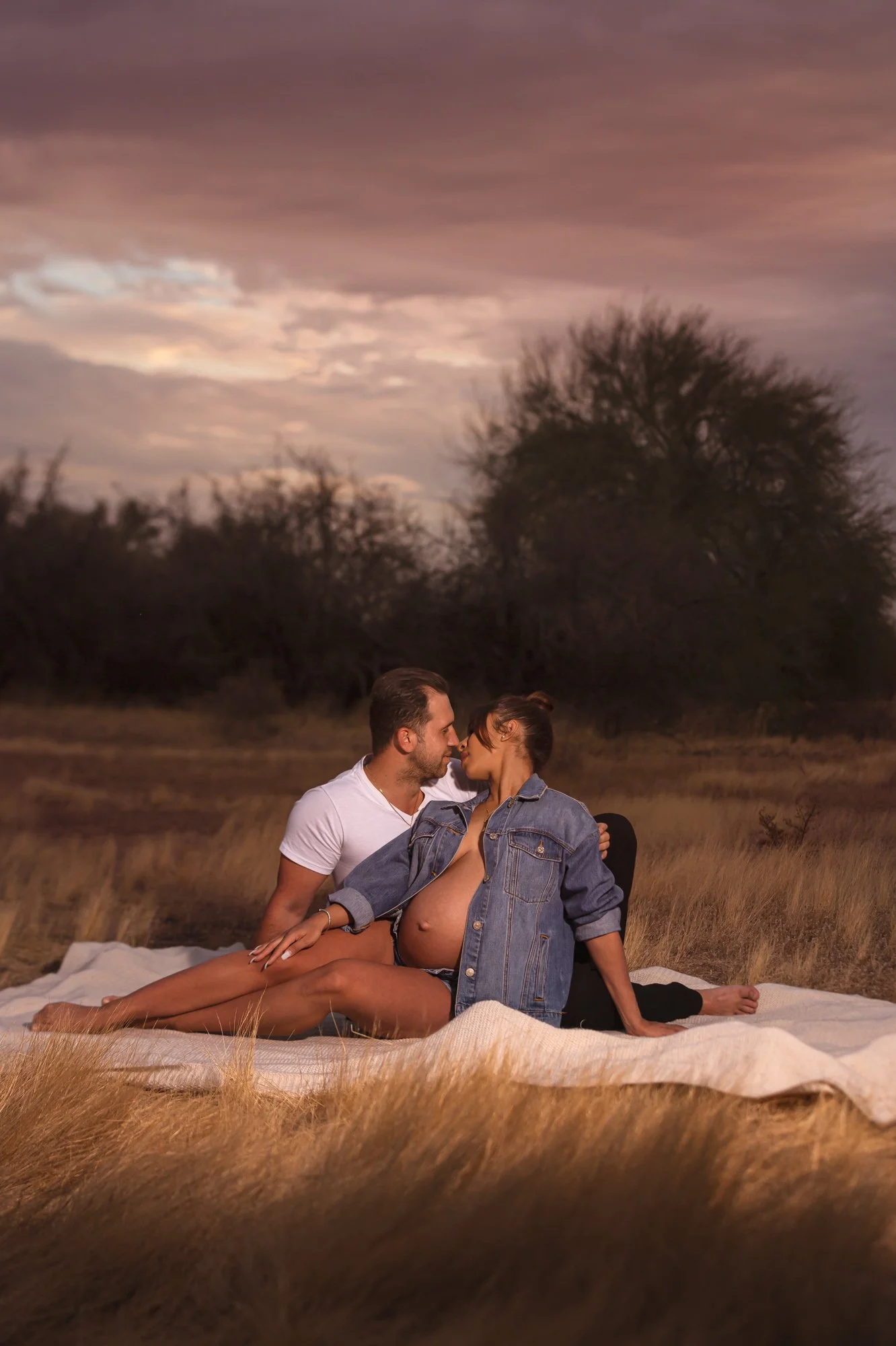 Pregnant couple sitting on a blanket in grassy field at sunset.