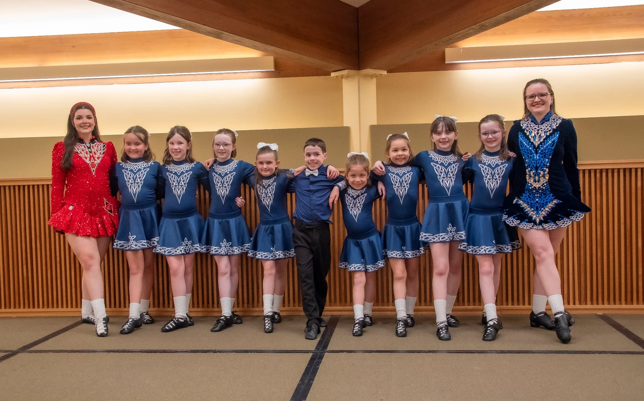 11 dancers stand in a row smiling at the camera. Most of them are wearing blue, white, and grey dresses, with one girl wearing a red dress and a boy in a blue shirt and navy pants.