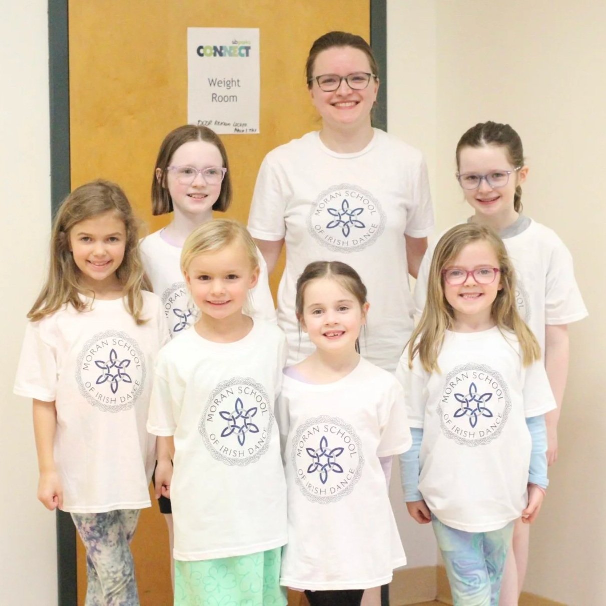 6 young girls stand with their teacher smiling at the camera. They are all in matching white shirts with the Moran School logo