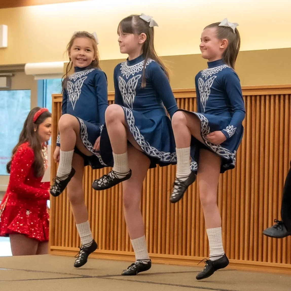 Three young girls in blue, white, and grey dresses smile as they dance with one foot pointed to their knee