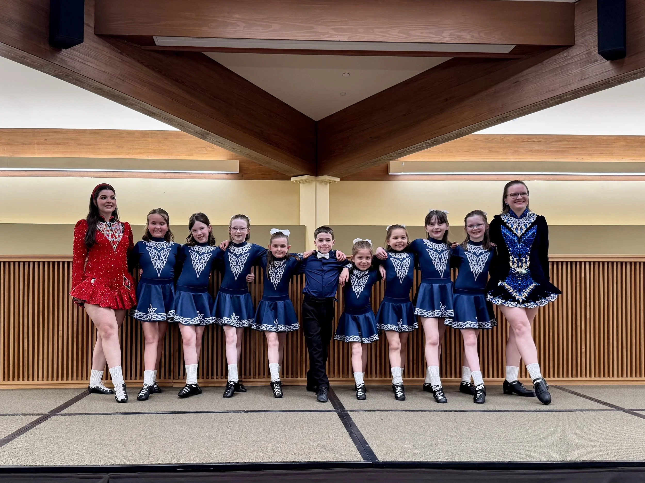 11 dancers stand on stage smiling with their toes pointed. Most of the dancers are in blue, white, and grey outfits, with one taller dancer wearing a red dress
