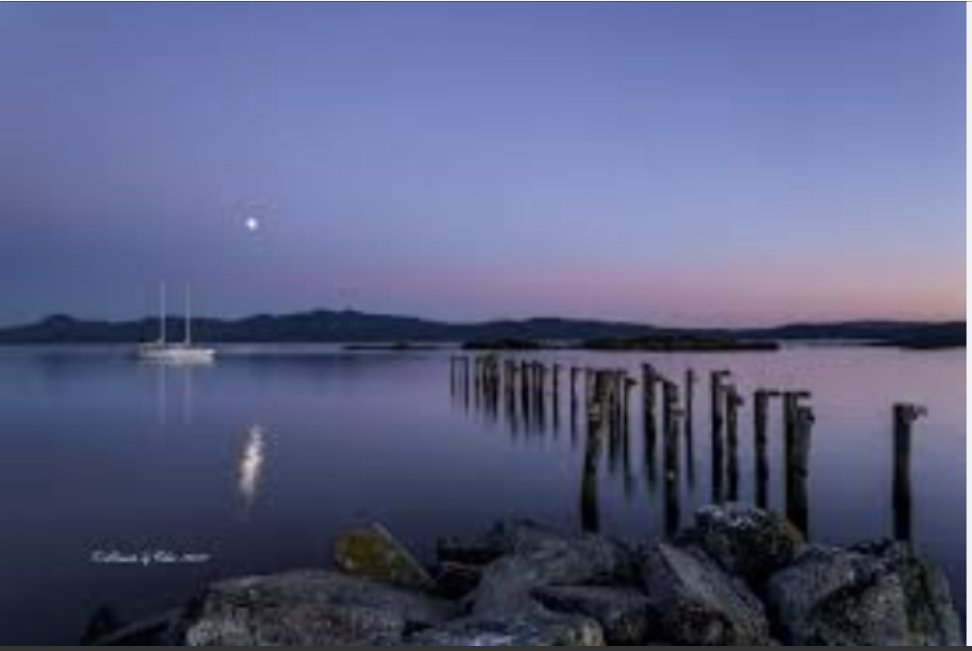 Tranquil dusk scene with a sailboat on a calm lake, moon and reflection visible, old wooden pilings extending into water, rocky shore in foreground, and distant hills on horizon.