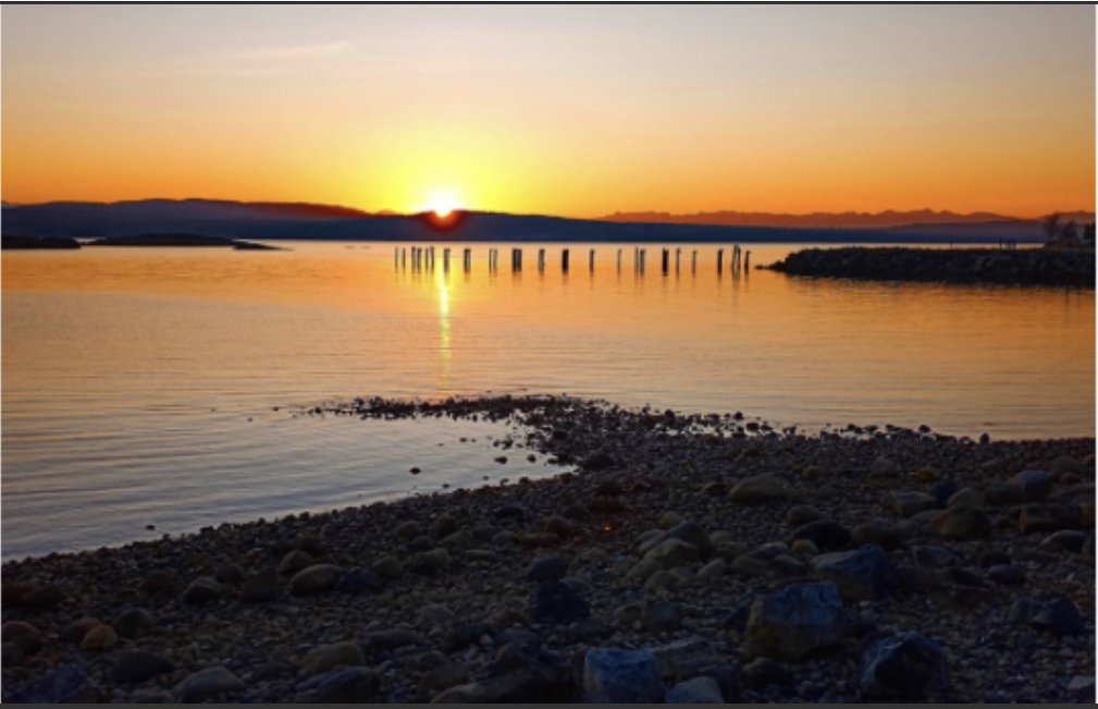 Sunset over calm water with a rocky shoreline, wooden posts, and distant mountains.