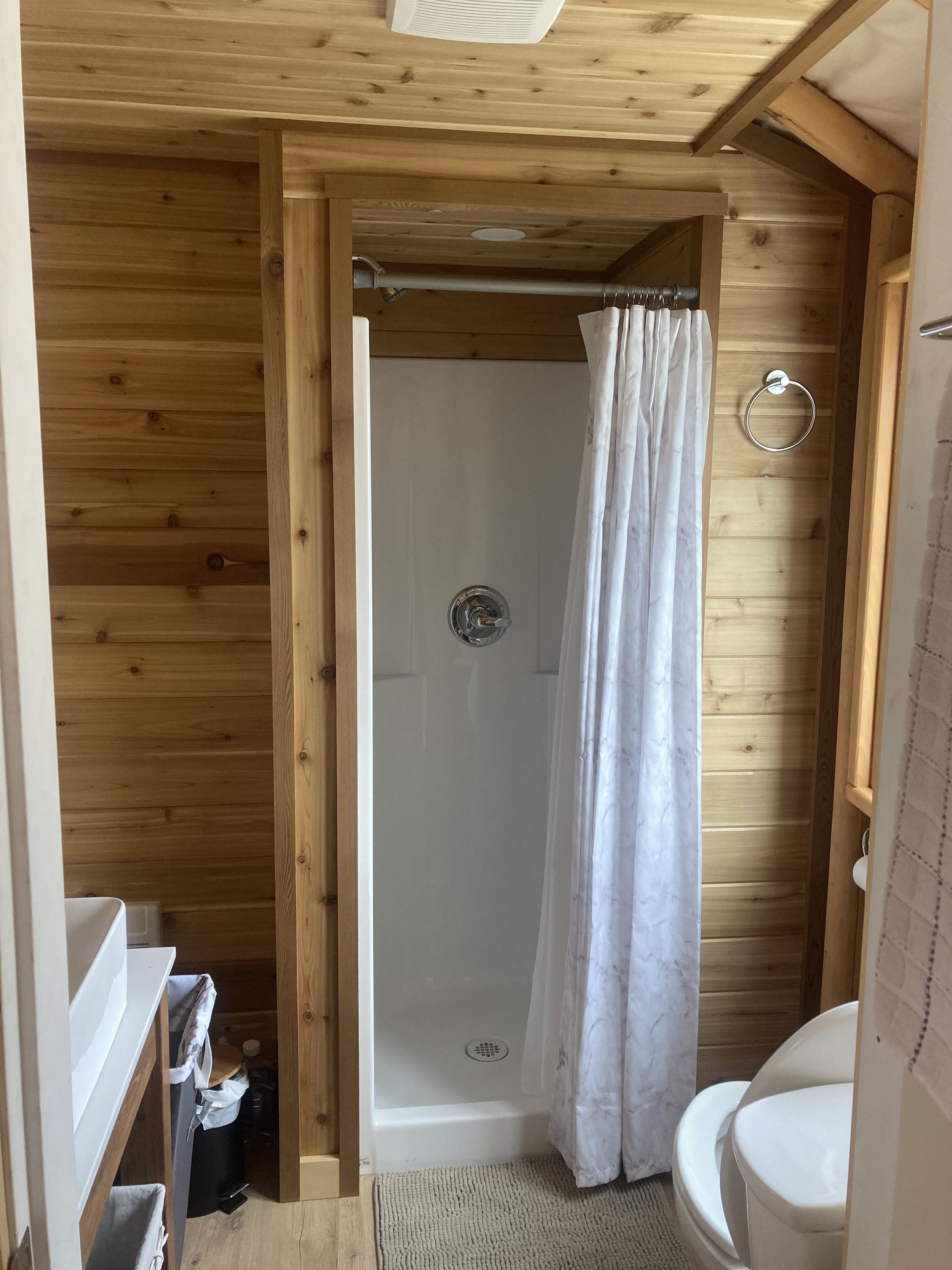 Small bathroom with wooden walls, shower stall with curtain, white toilet, sink, and wall-mounted holder.