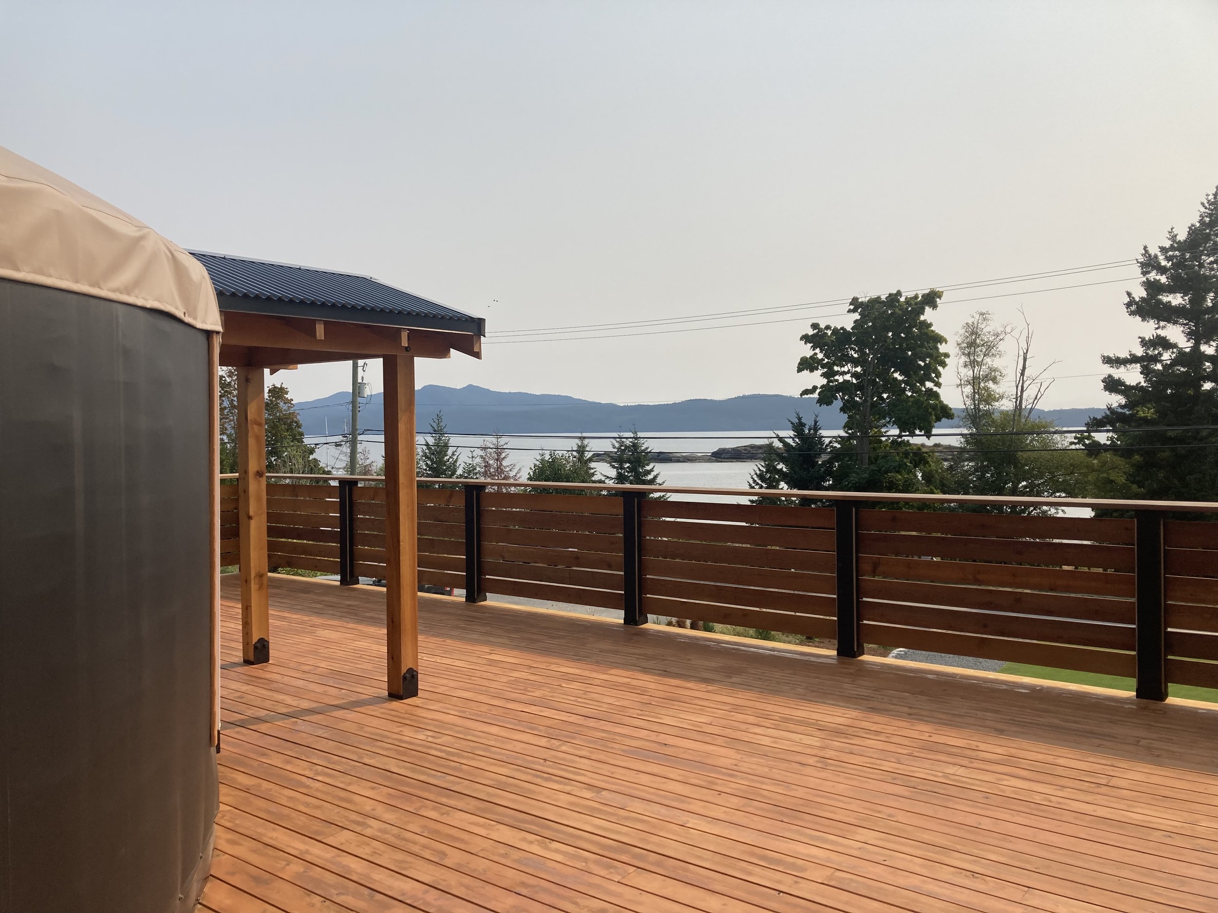 Wooden deck with railing overlooking a body of water and distant hills, partial view of a yurt and a small wooden structure with a roof.