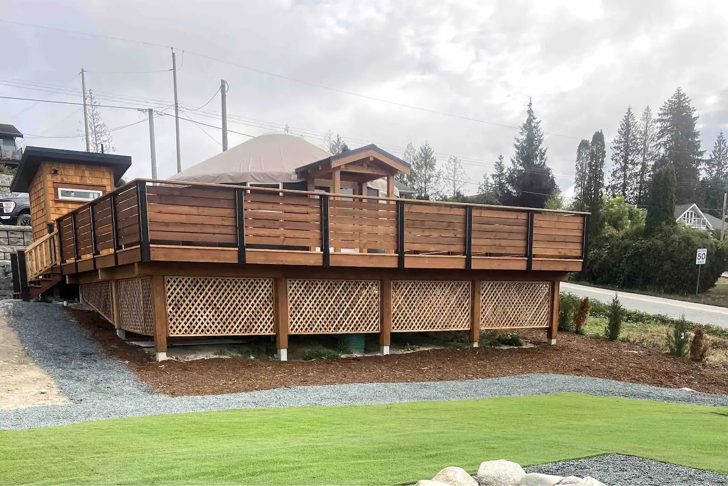 Wooden deck with lattice privacy screen, gravel path, and grassy area in front, surrounded by trees and power lines.