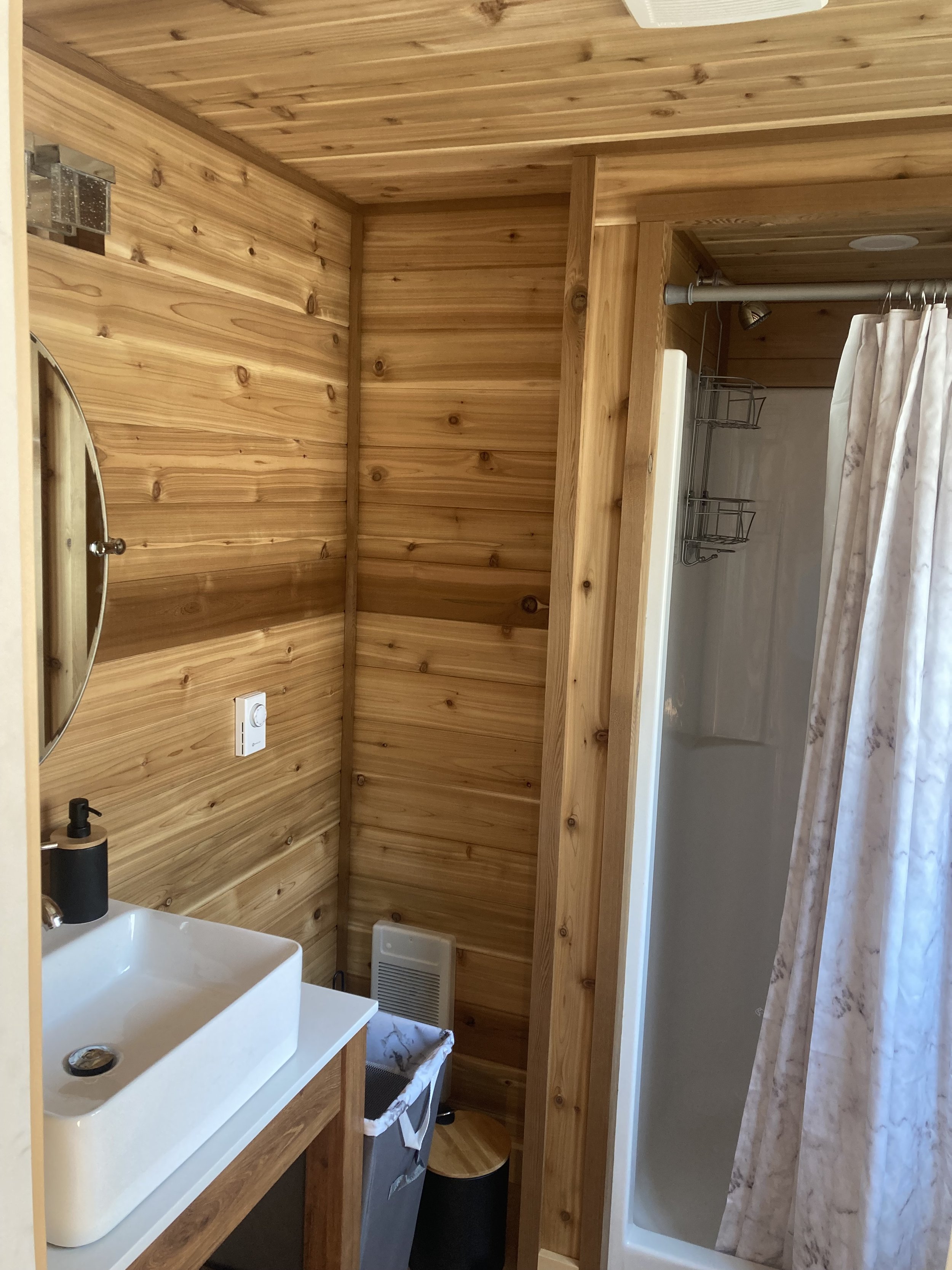 Small wooden bathroom with a rectangular sink, round mirror, and a shower stall with a curtain.