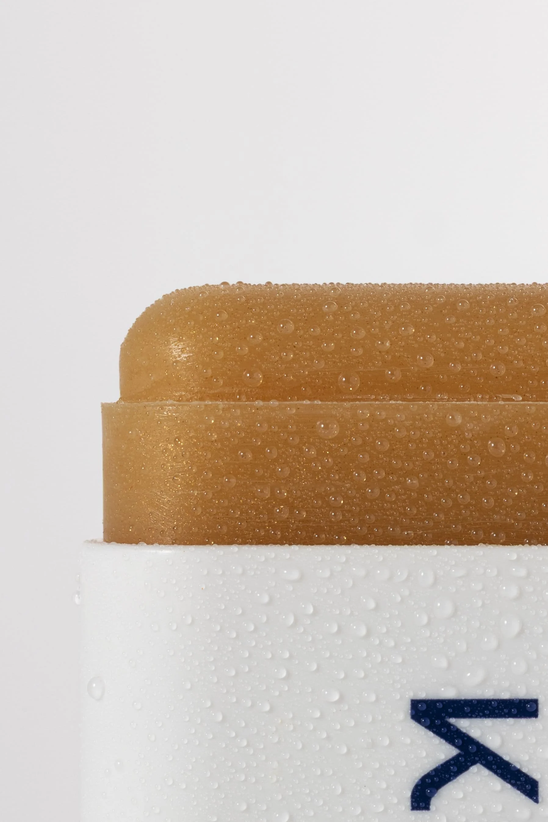 Une barre de savon beige avec des gouttelettes d'eau, posée sur un contenant blanc avec des gouttelettes d'eau et une partie d'une lettre bleue visible.