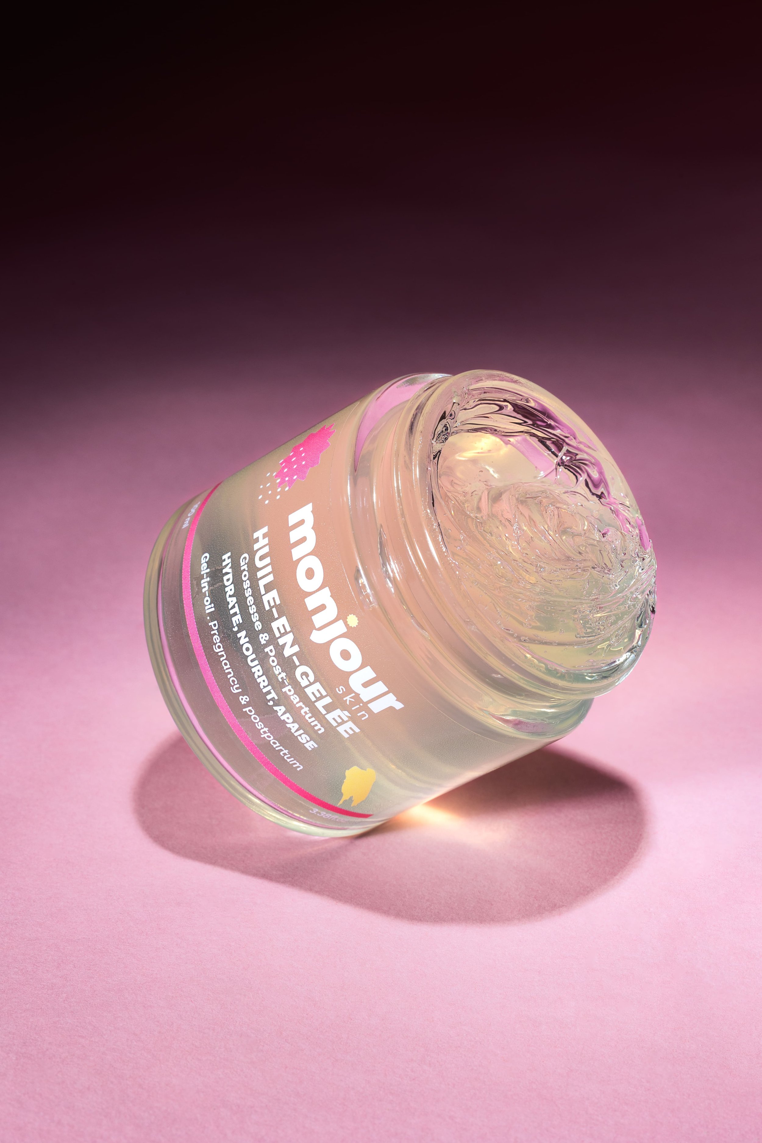 Pot de gelée transparent avec étiquette Monjour, posé sur un fond rose, créant une ombre port-à-port. La gelée semble être un produit de soin huilé, sans parfum, pour grossesse et post-partum.
