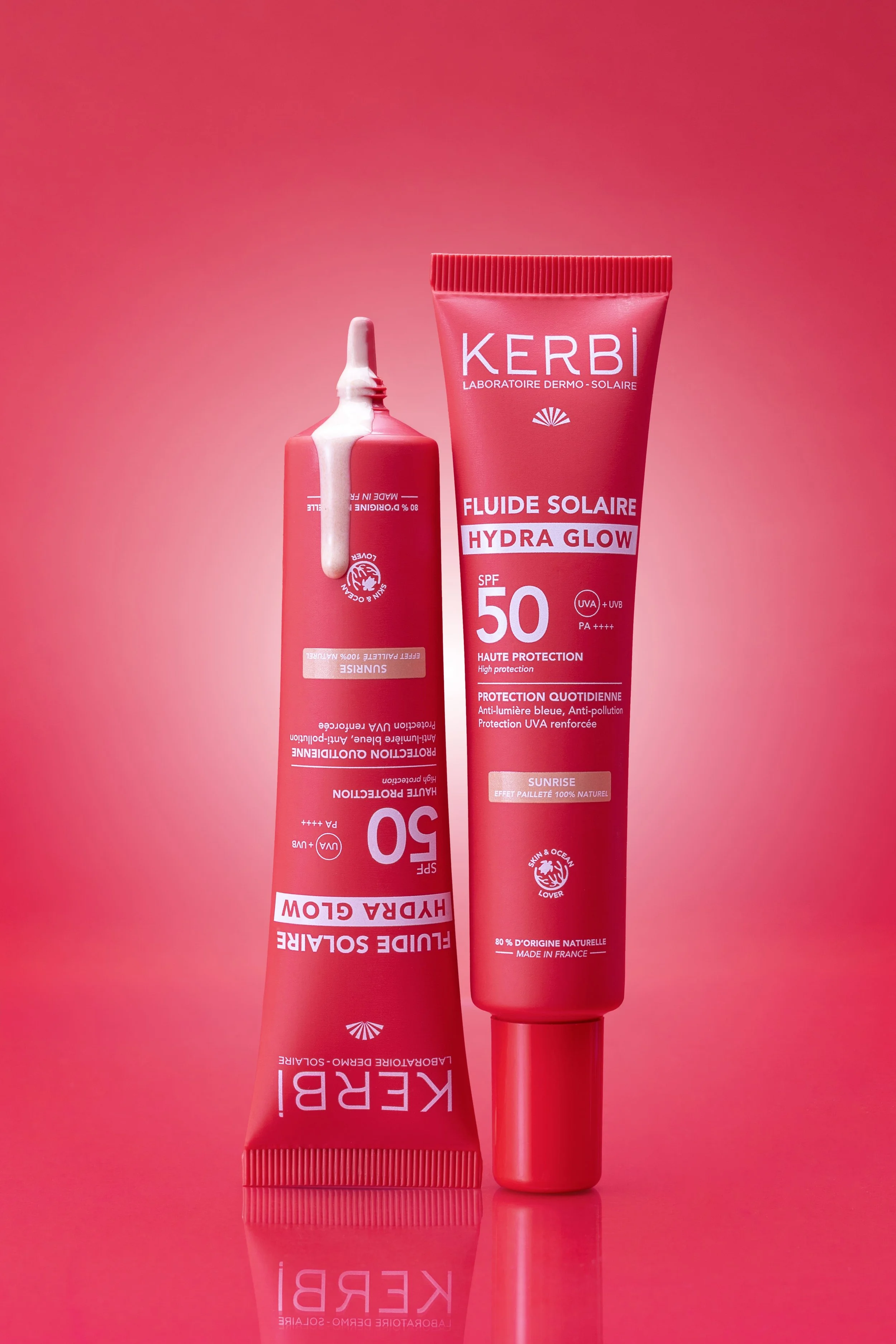 Tube de crème solaire Kerbi Hydra Glow SPF 50 posé à côté d'une autre tube de crème solaire, tous deux sur un fond rose avec réflexion