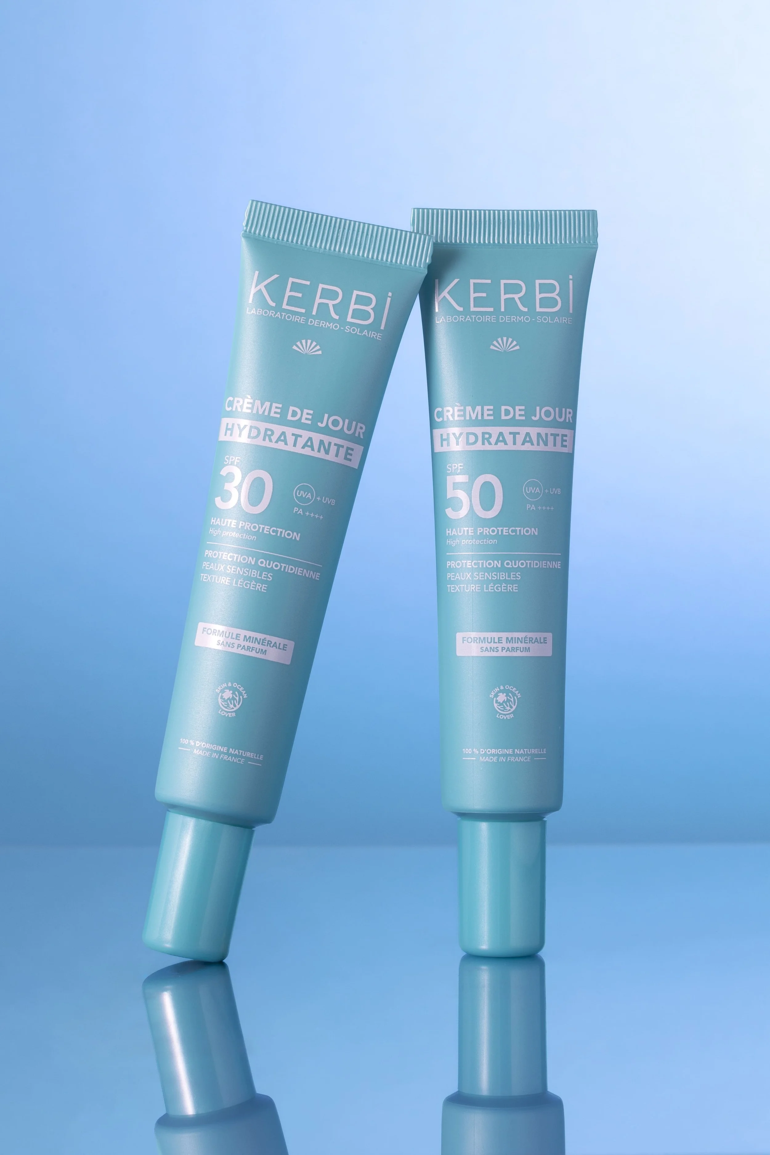 Deux tubes de crème solaire de marque Kerbi, un avec SPF 30 et l'autre SPF 50, positionnés verticalement sur un fond bleu, leur reflet visible en dessous.