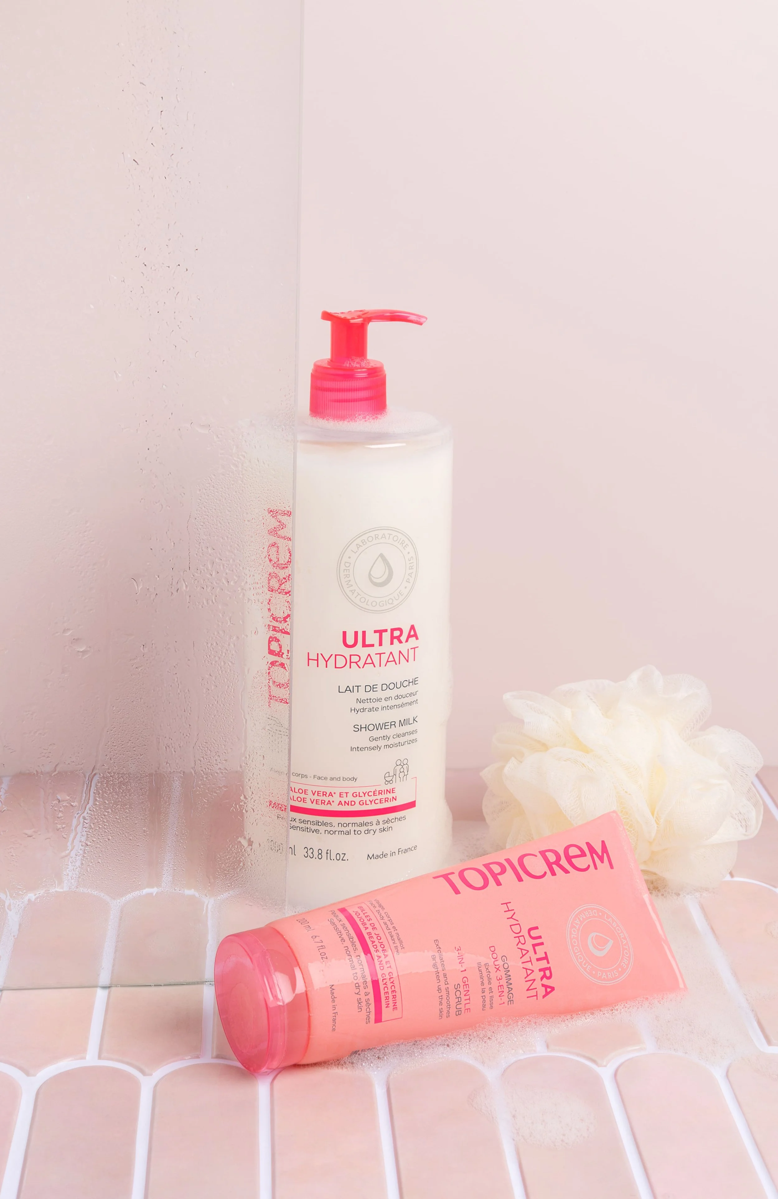 Produits de soins pour la peau et le corps, notamment un flacon de lait de douche et un tube de gommage hydratant Ultra, avec une éponge de bain, sur une surface en carreaux de céramique.