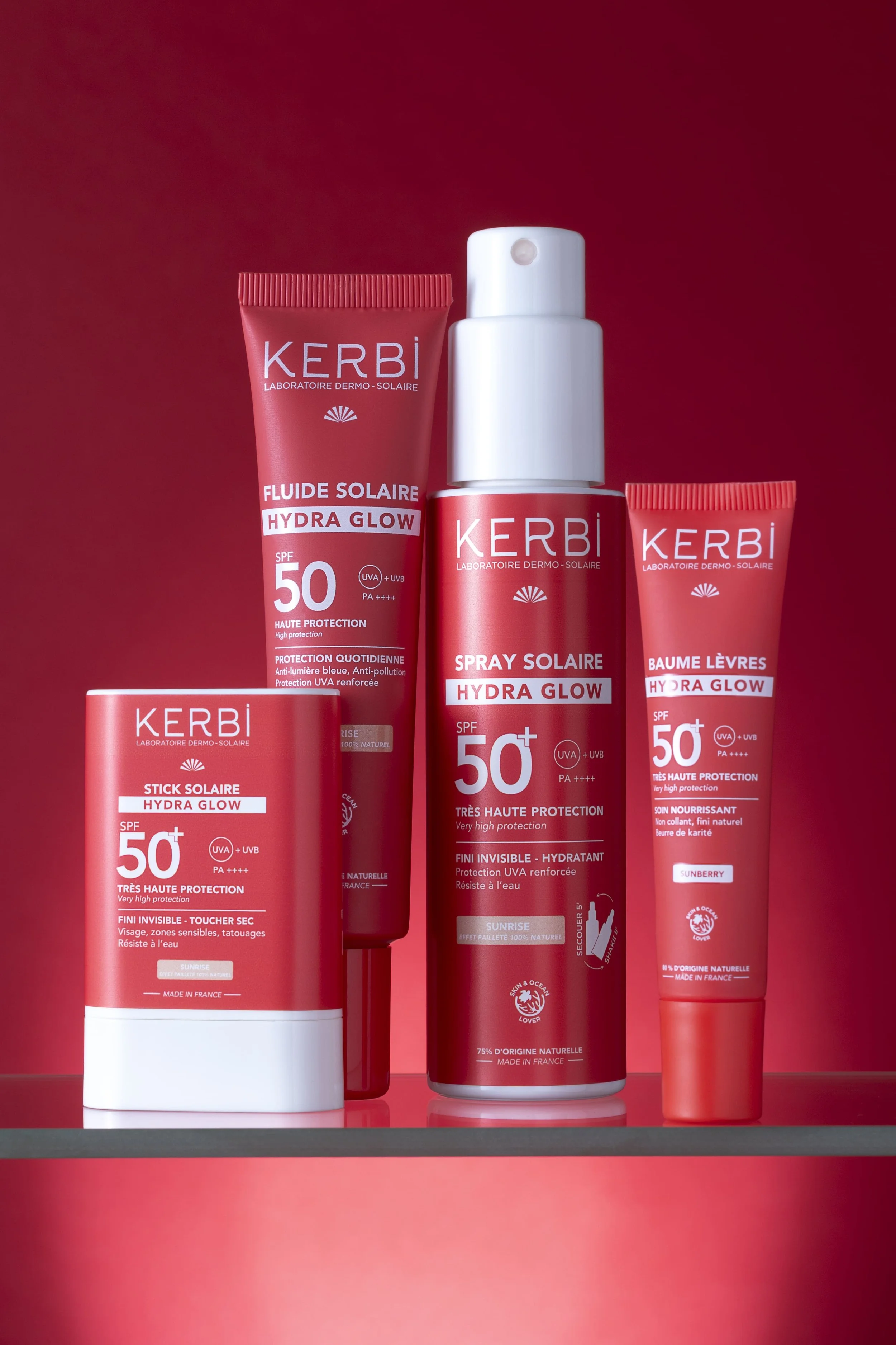 Gamme de produits solaires Kerbi, incluant un fluide solaire Hydra Glow SPF 50, un spray solaire Hydra Glow SPF 50, un stick solaire Hydra Glow SPF 50, et un baume lèvres Hydra Glow SPF 50, tous présentés sur une surface contre un fond rouge.