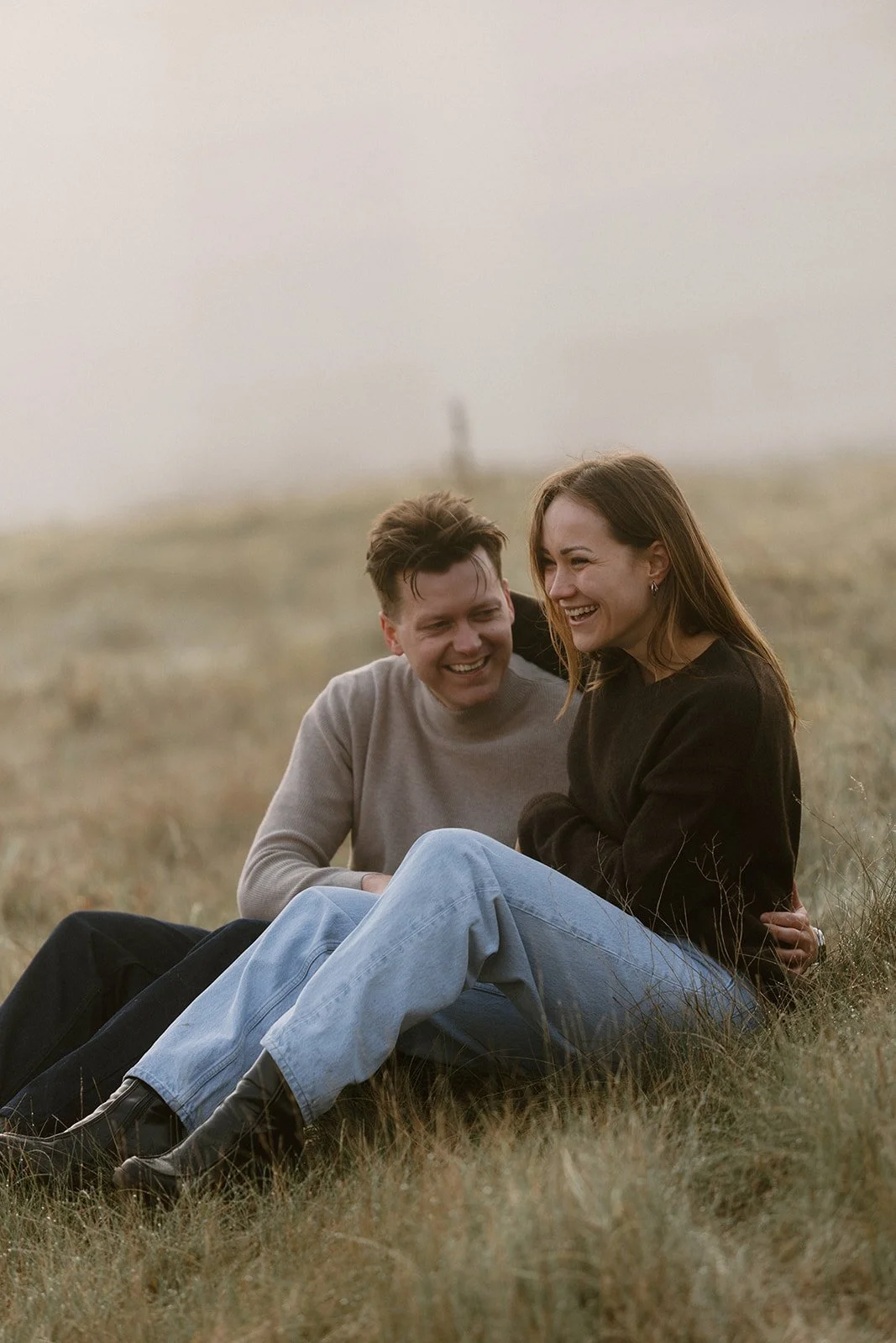 A young man and woman sitting on grass outdoors, laughing and smiling, enjoying each other's company.
