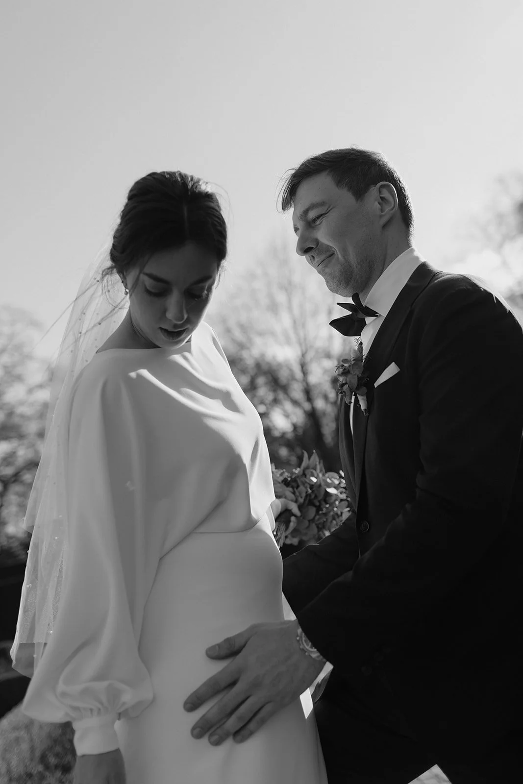 Black and white photo of a bride and groom during their wedding ceremony outdoors. The bride is looking down at her pregnant belly with a surprised expression, and the groom is smiling looking at her, holding her belly with one hand.