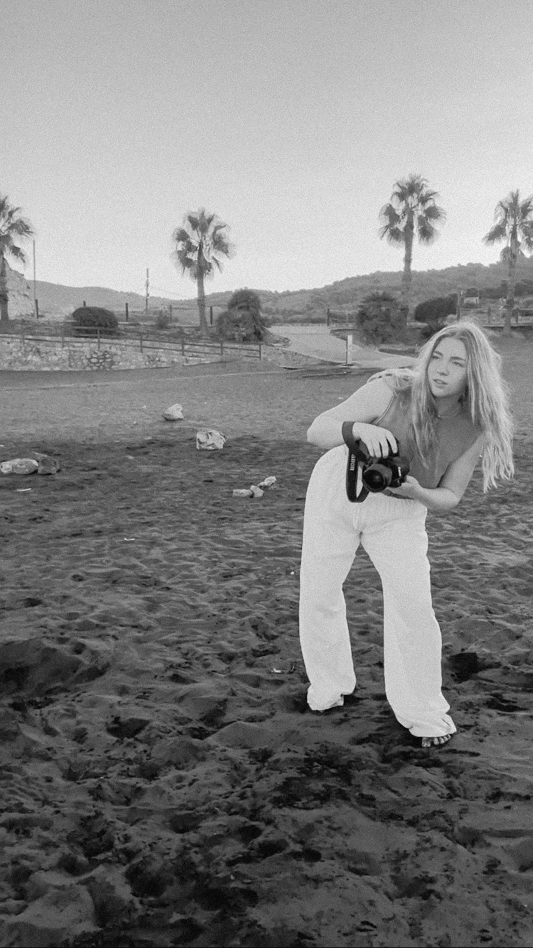 A young woman with long hair holding a camera in a sandy outdoor area with palm trees and hills in the background.