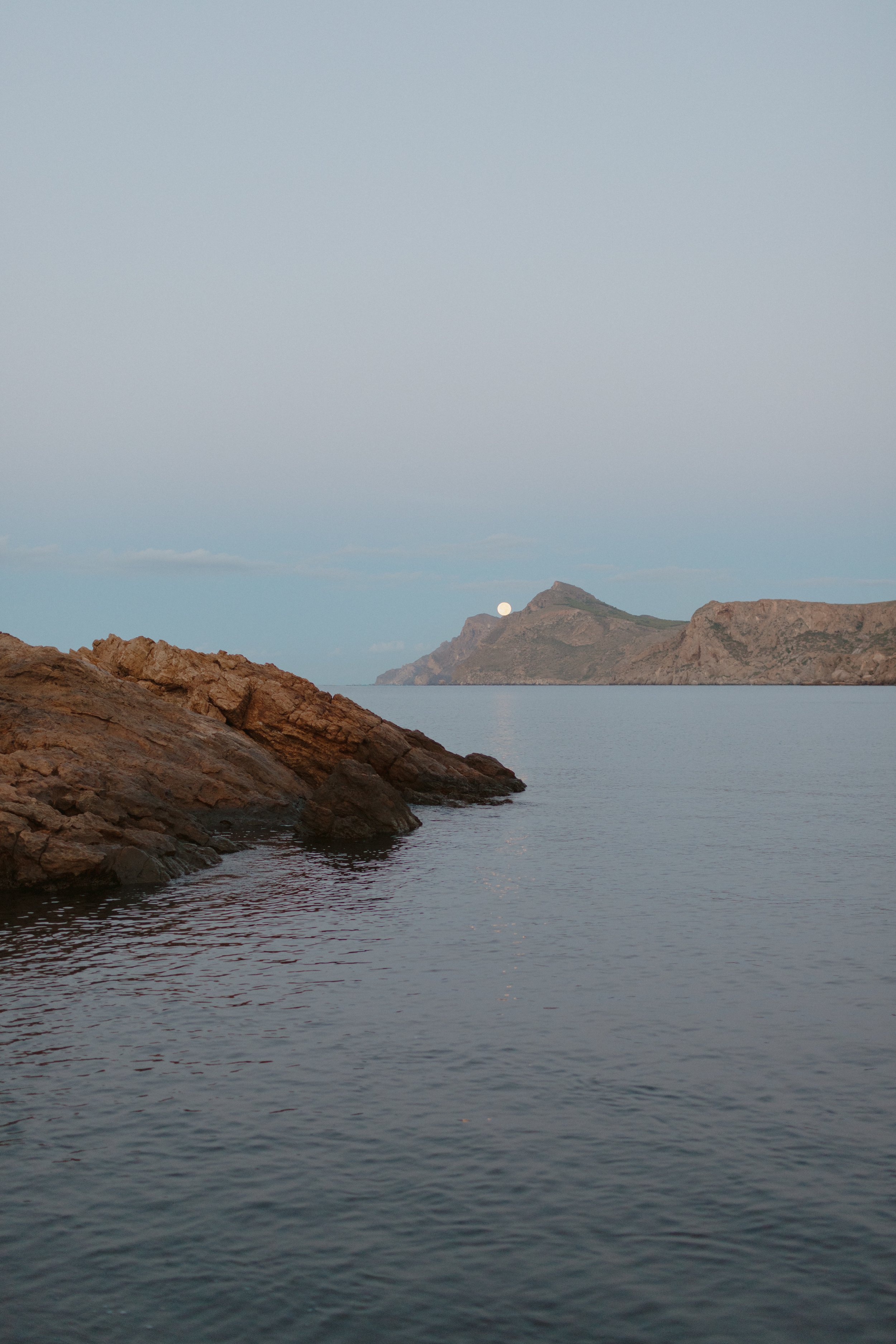 A serene coastal scene with rocky shoreline on the left, calm water, and distant landmass under a pale sky with the moon visible near the horizon.