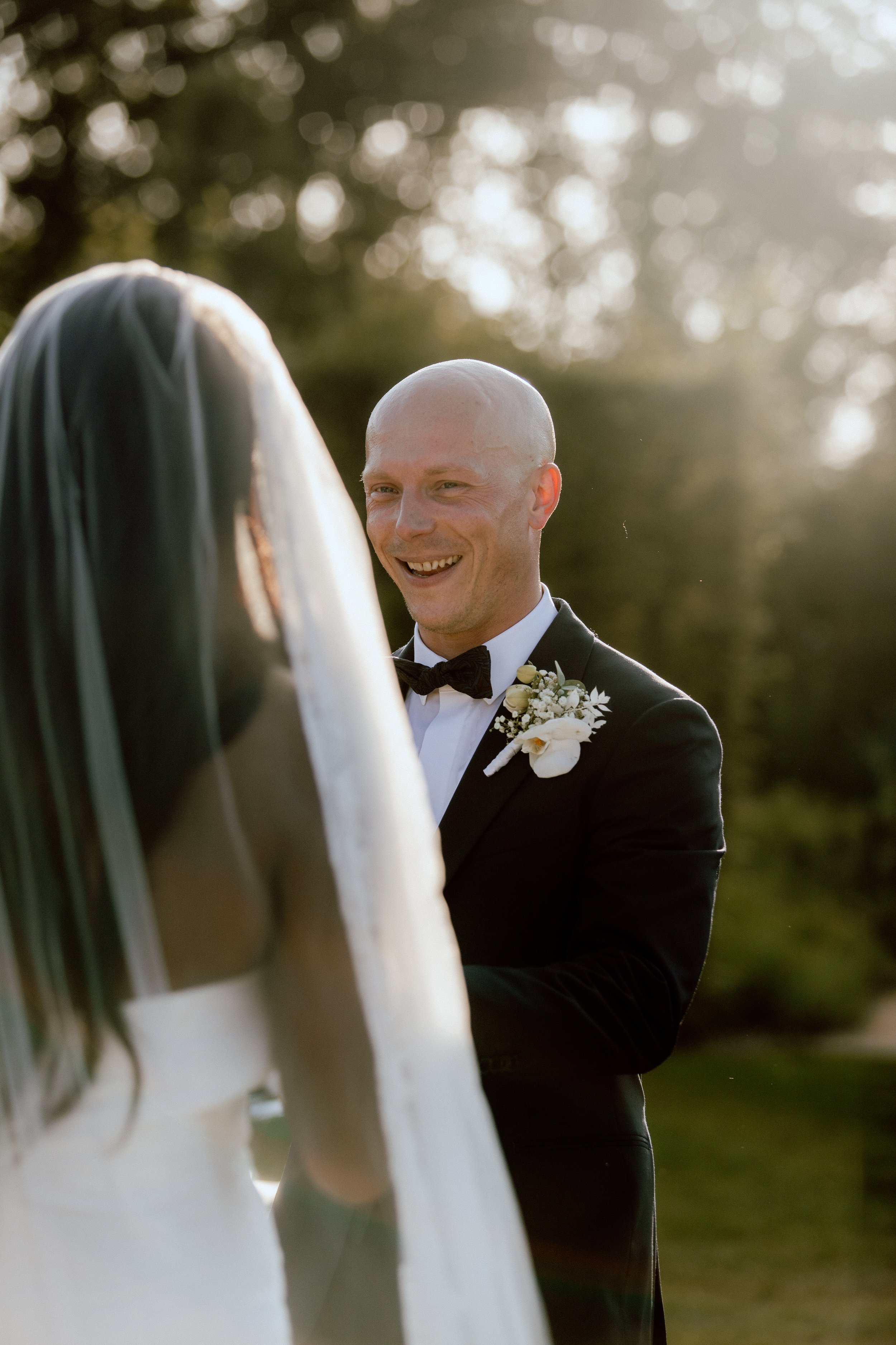 A groom with a bald head and black tuxedo with a bow tie, smiling at his bride during an outdoor wedding. The bride, with long dark hair and veil, is partially visible and facing the groom. The background shows blurred trees and sunlight.