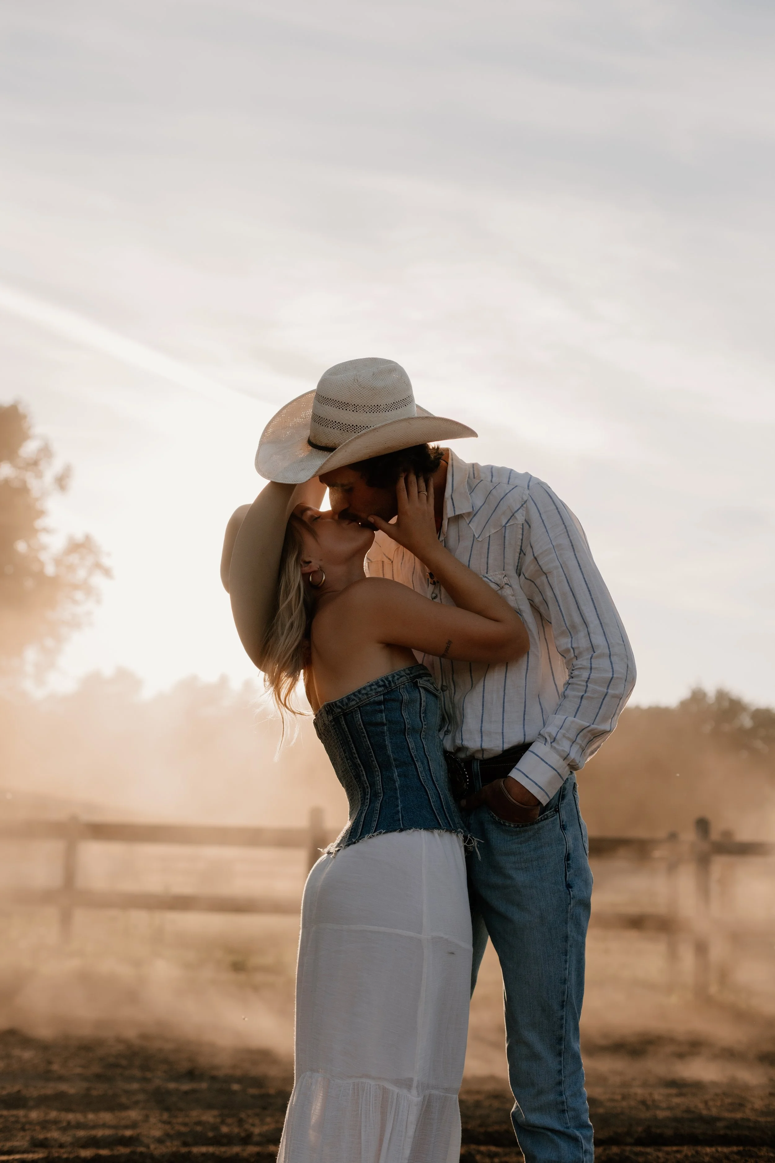A couple sharing a kiss outdoors at sunset, with dusty air and a fence in the background.