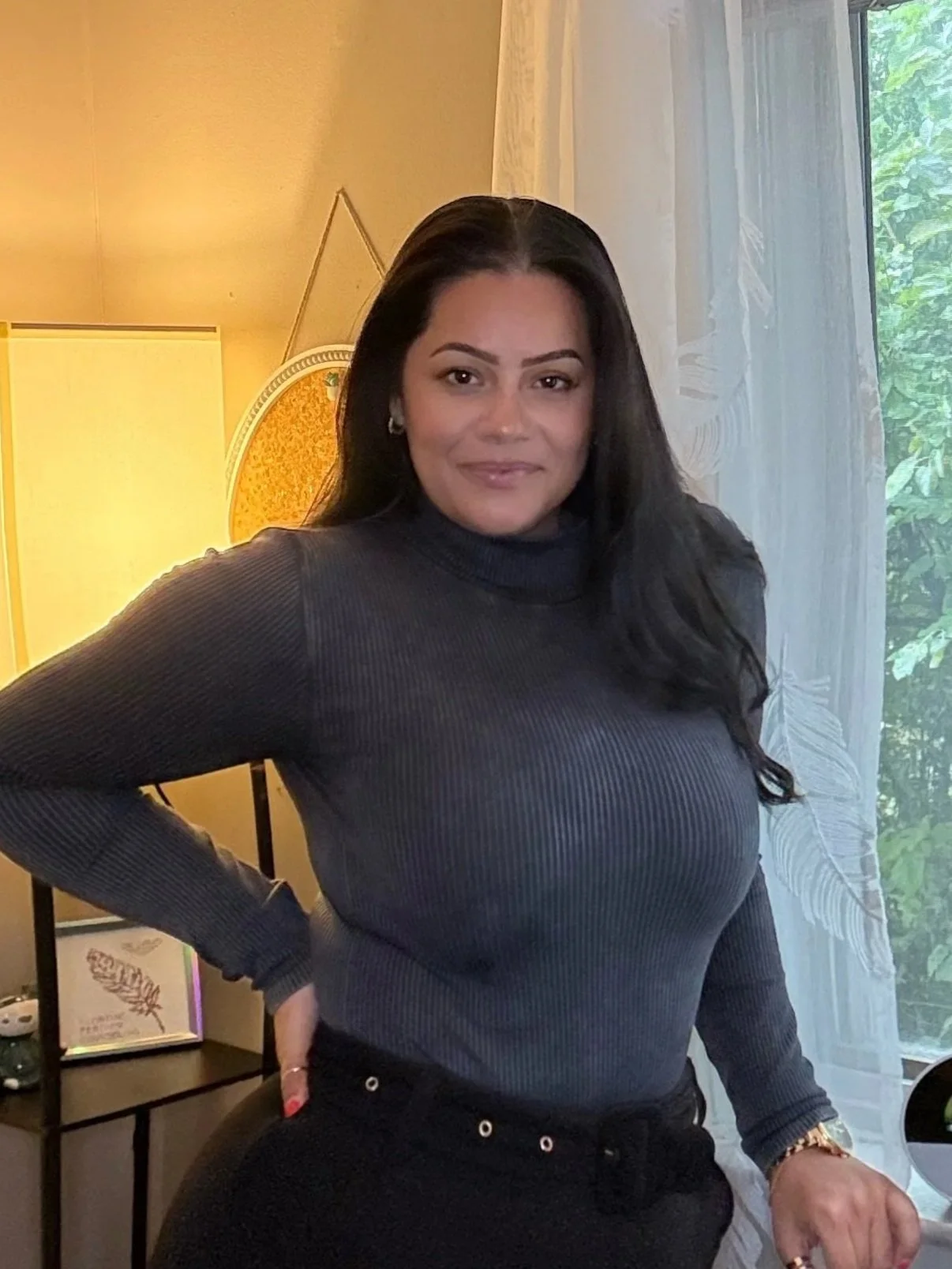 A woman with long black hair, wearing a dark gray turtleneck, standing indoors next to a window with white curtains and greenery outside. She is smiling slightly, with one hand on her hip and the other resting on a surface.