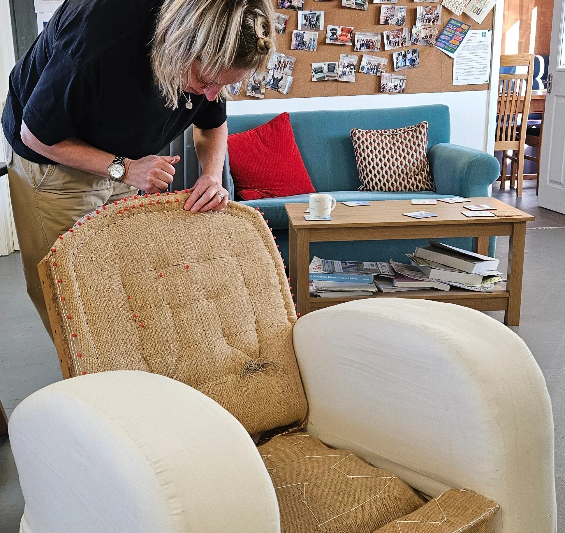 A woman working on reupholstering a beige armchair with pins and fabric in a cozy living room. The room has a teal sofa with red and patterned pillows, a coffee table with papers and a mug, and a bulletin board with photos on the wall.