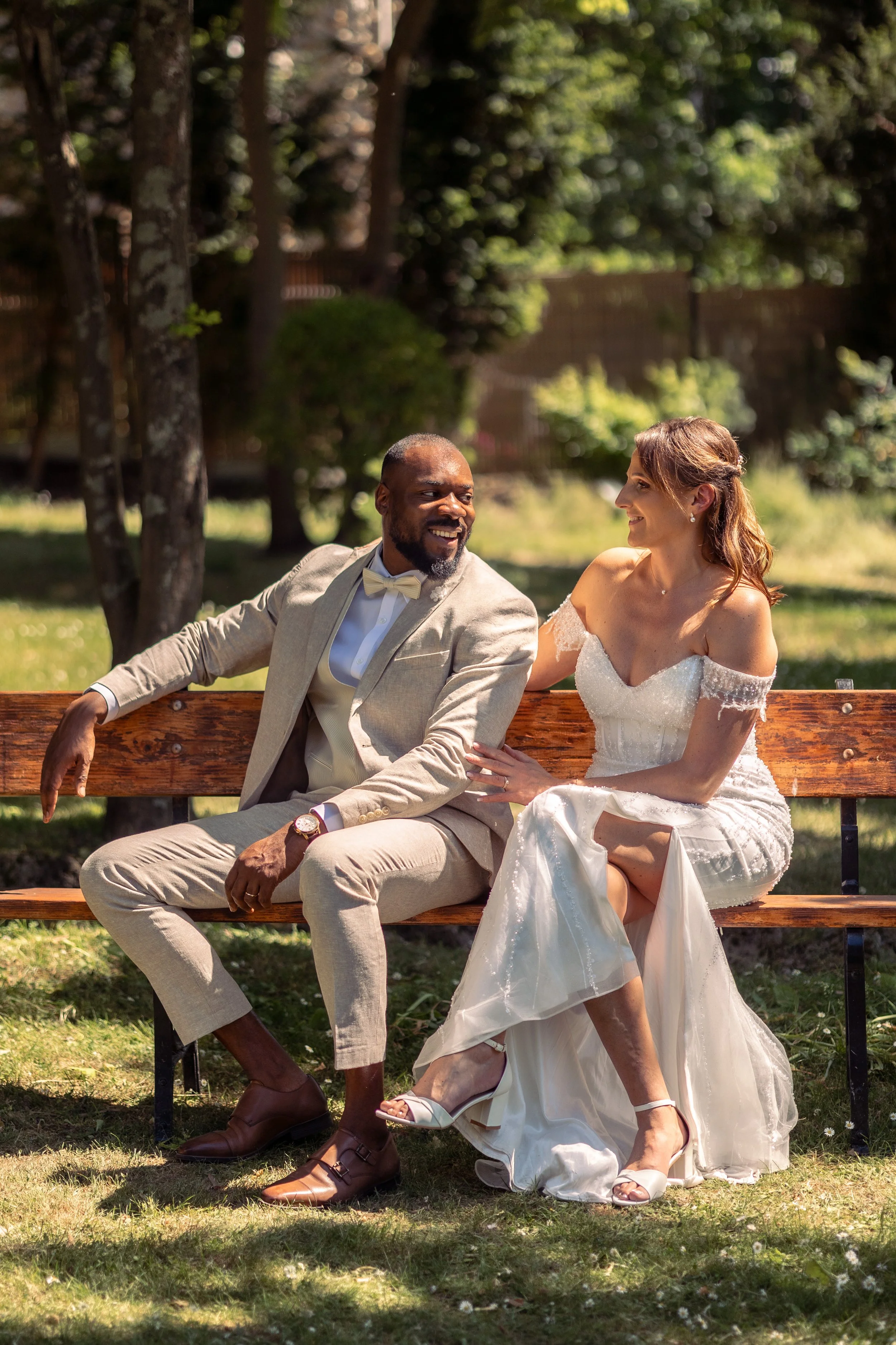 Un couple souriant assis sur un banc en arrière-plan d'un parc, lors d'un mariage. La femme porte une robe de mariée blanche, et l'homme un costume beige avec nœud papillon.