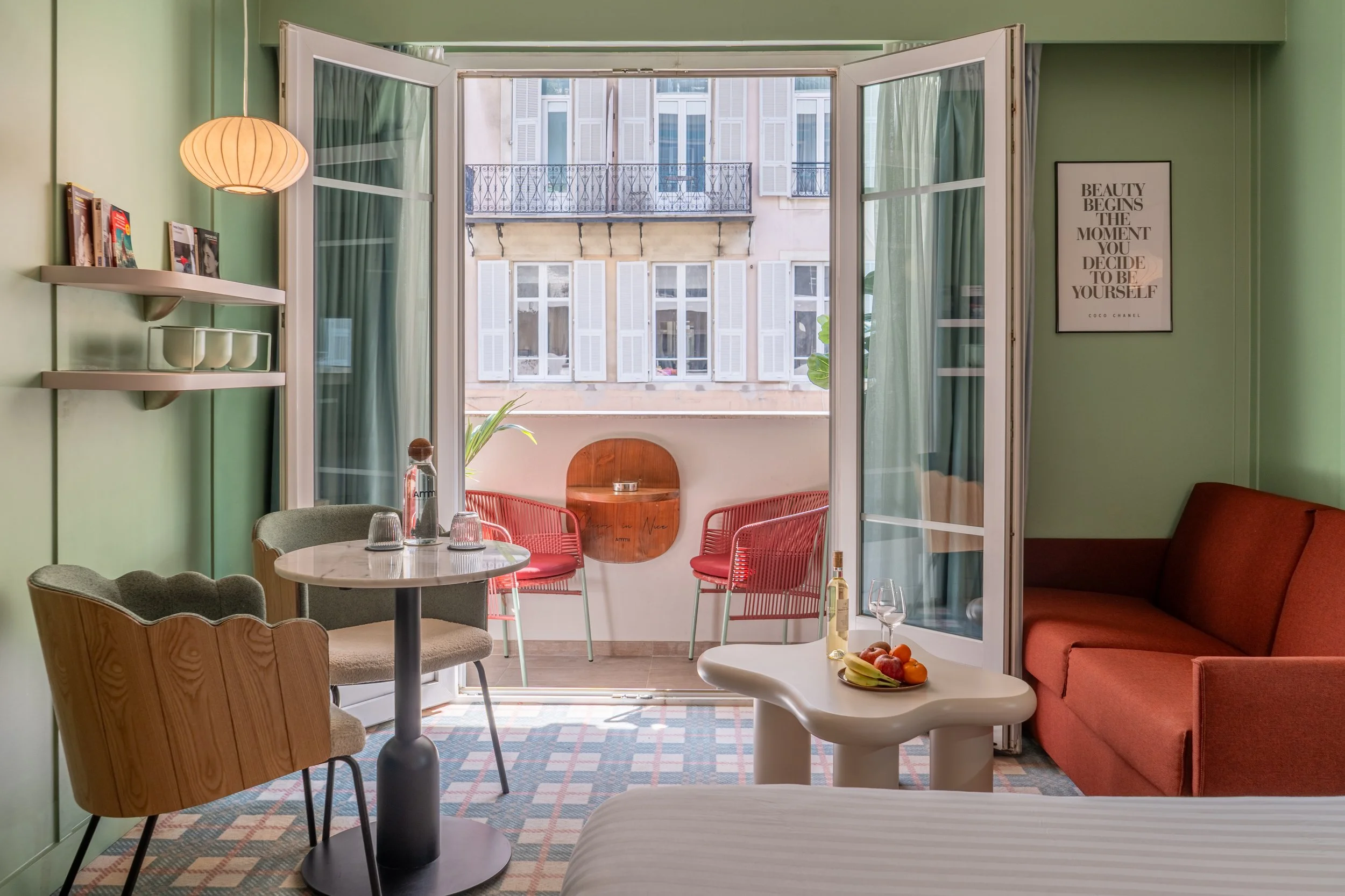 Salon avec vue sur un balcon. Murs verts, canapé rouge, table blanche avec fruits, assiette, et verre de vin, chaise en bois et en tissu, étagère avec magazines, décoration minimaliste, fenêtre grande ouverte avec vue sur un immeuble parisien.