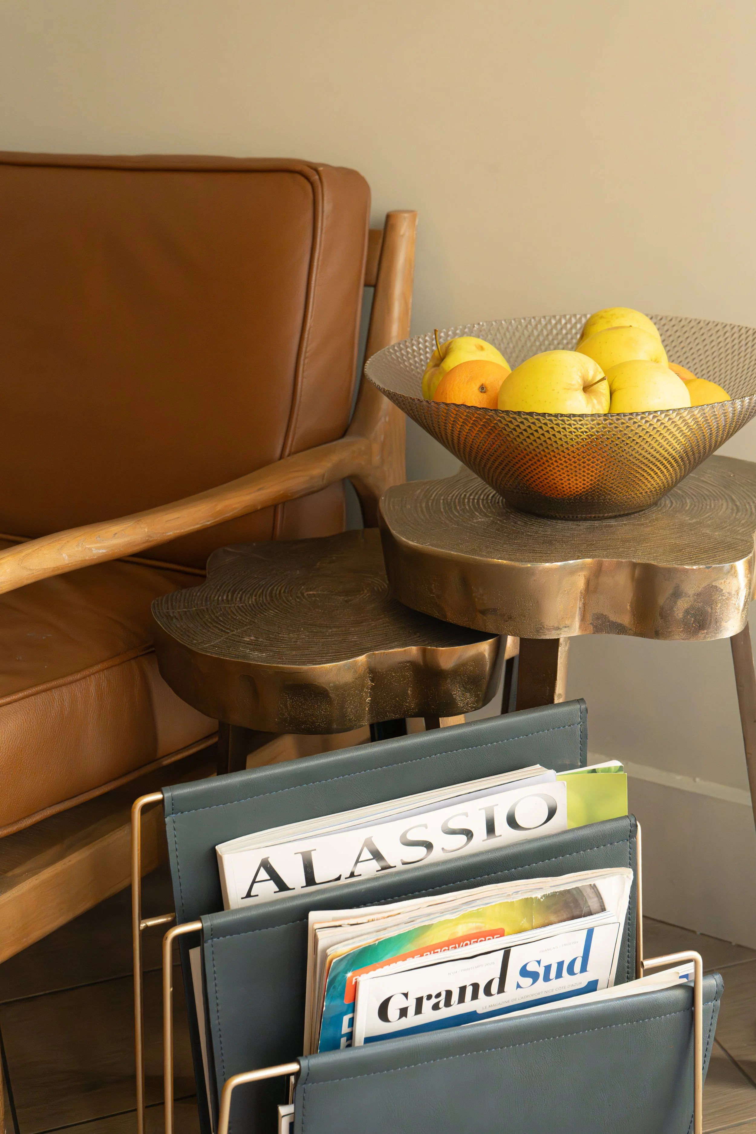 Une table en bois avec un bol contenant des pommes jaunes et oranges, un fauteuil en cuir brun, un porte-magazines avec des magazines en français, et deux petits tabourets en bois.