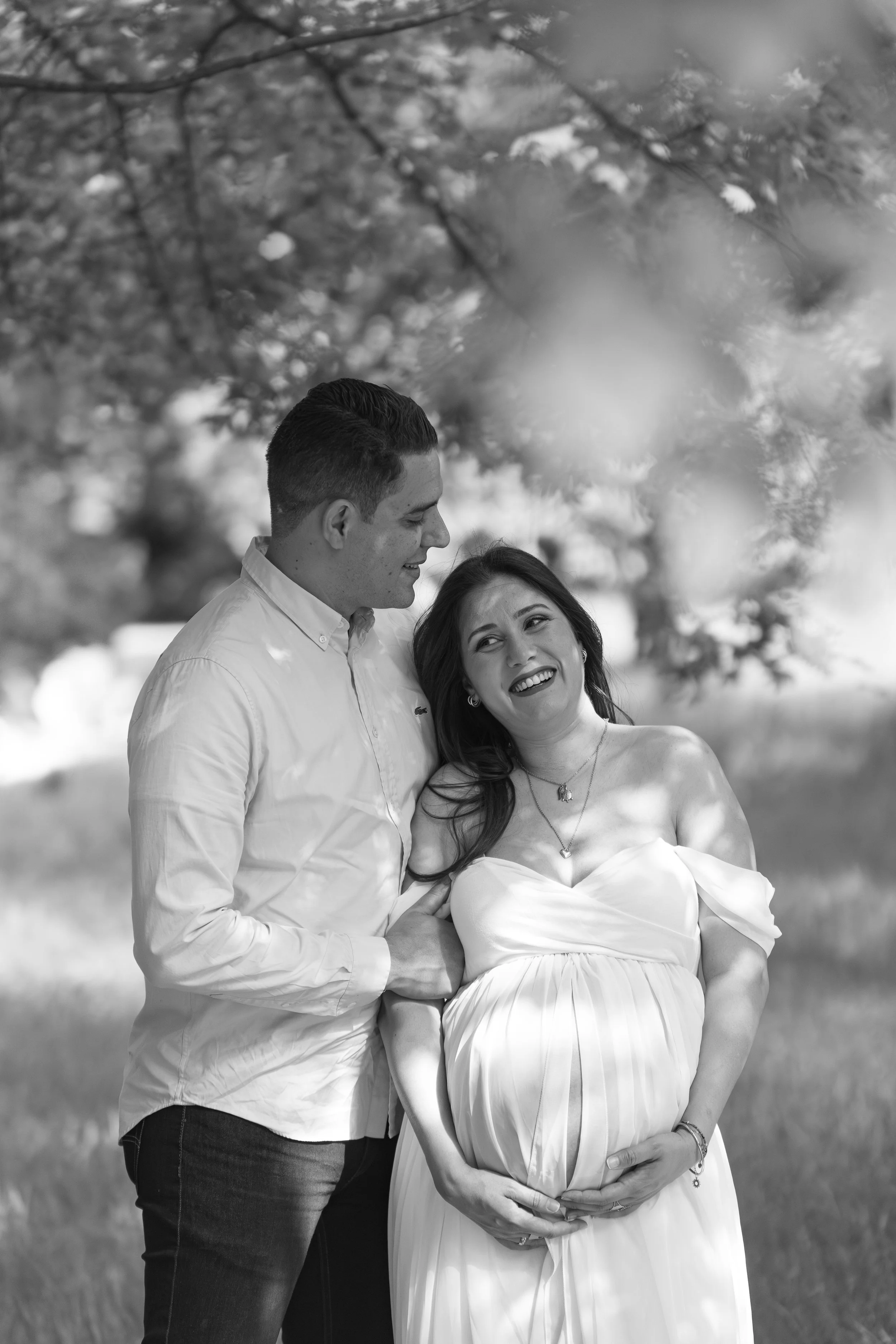 Une femme enceinte en robe blanche souriante, accompagnée d'un homme en chemise blanche, se tenant la main sous un arbre en plein air, en noir et blanc.
