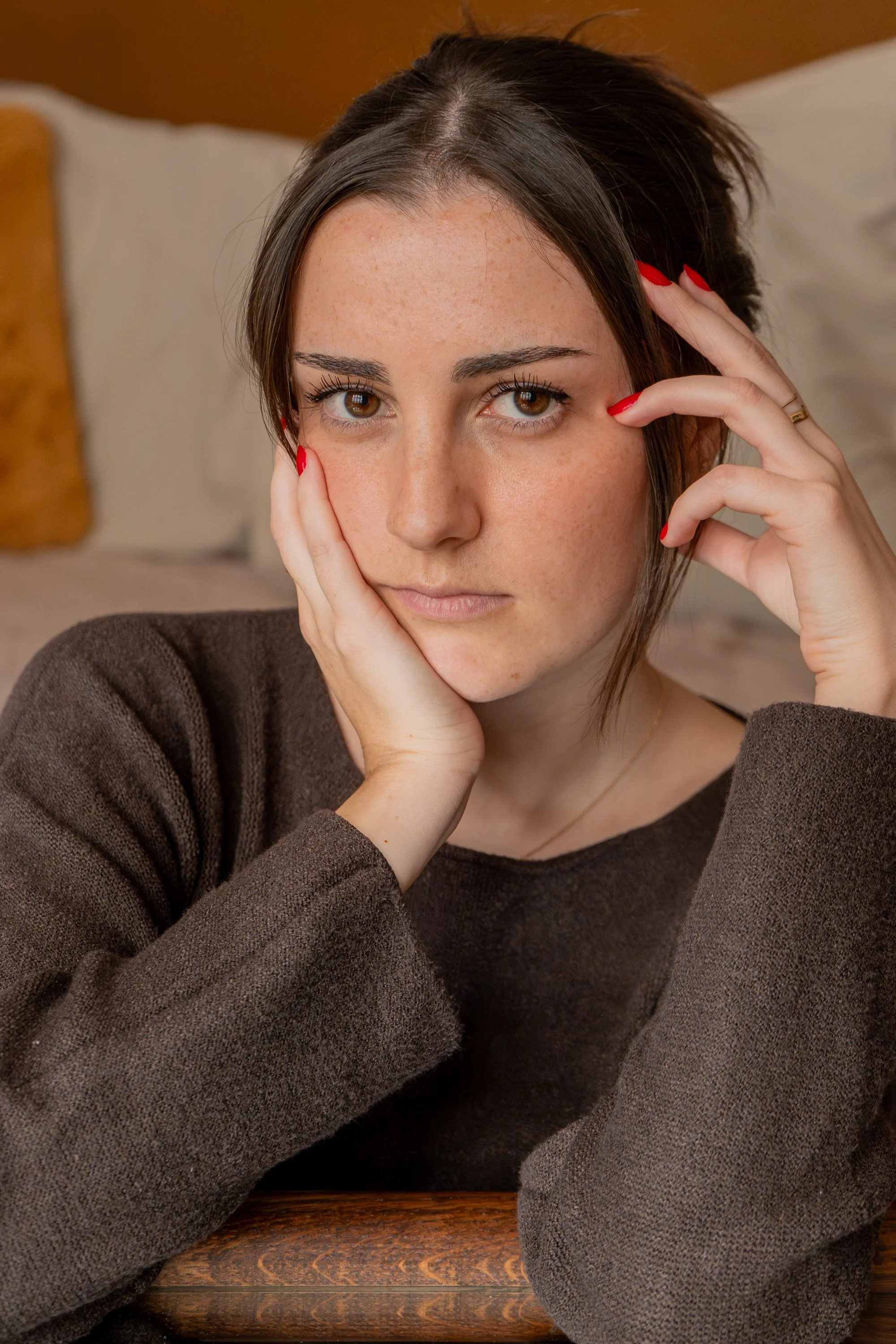 Jeune femme posant avec sa main sur sa joue, regardant vers l'objectif, avec des cheveux bruns, portant un pull marron, dans un intérieur chaleureux.