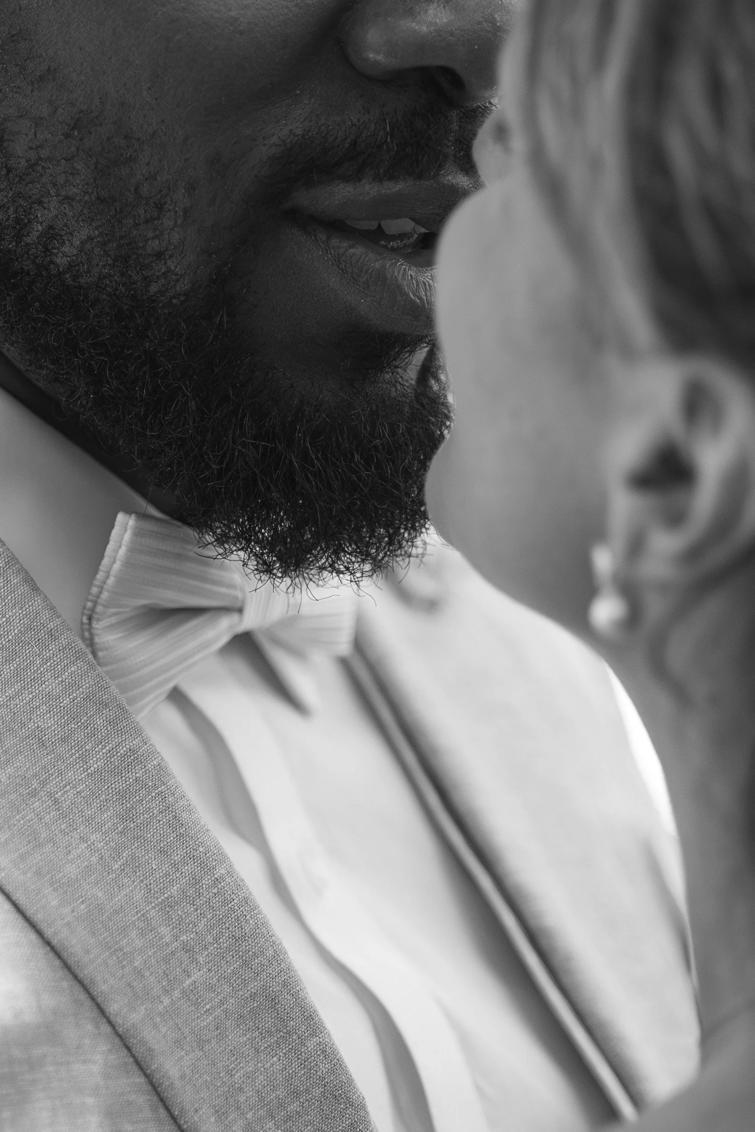 Gros plan d'un homme avec une barbe, portant un costume et une nœud papillon, se rapprochant d'une femme avec des cheveux courts, dans une scène en noir et blanc.