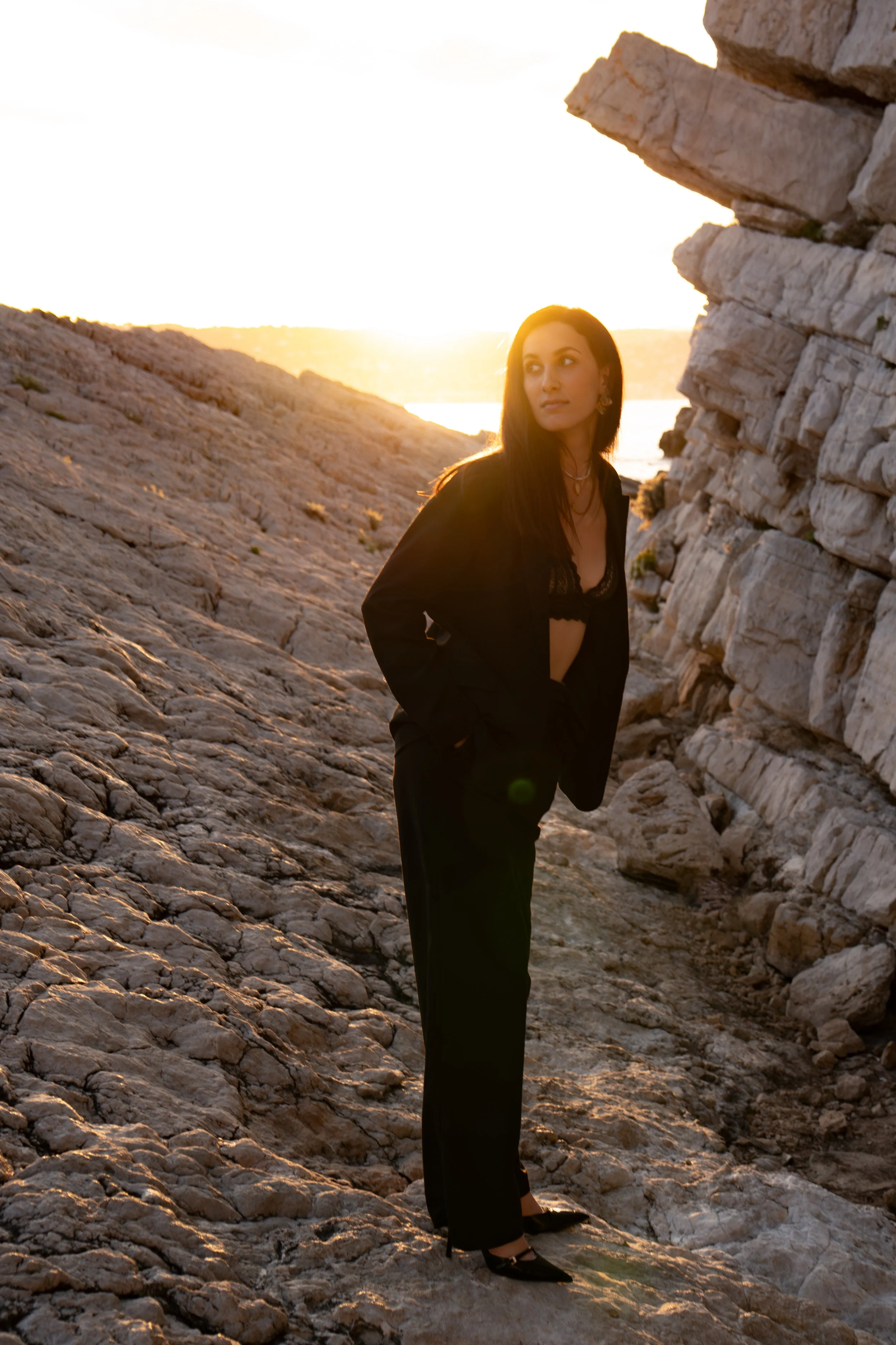 Femme en costume noir et lingerie, debout sur un terrain rocheux lors du coucher du soleil, près d'une formation rocheuse.