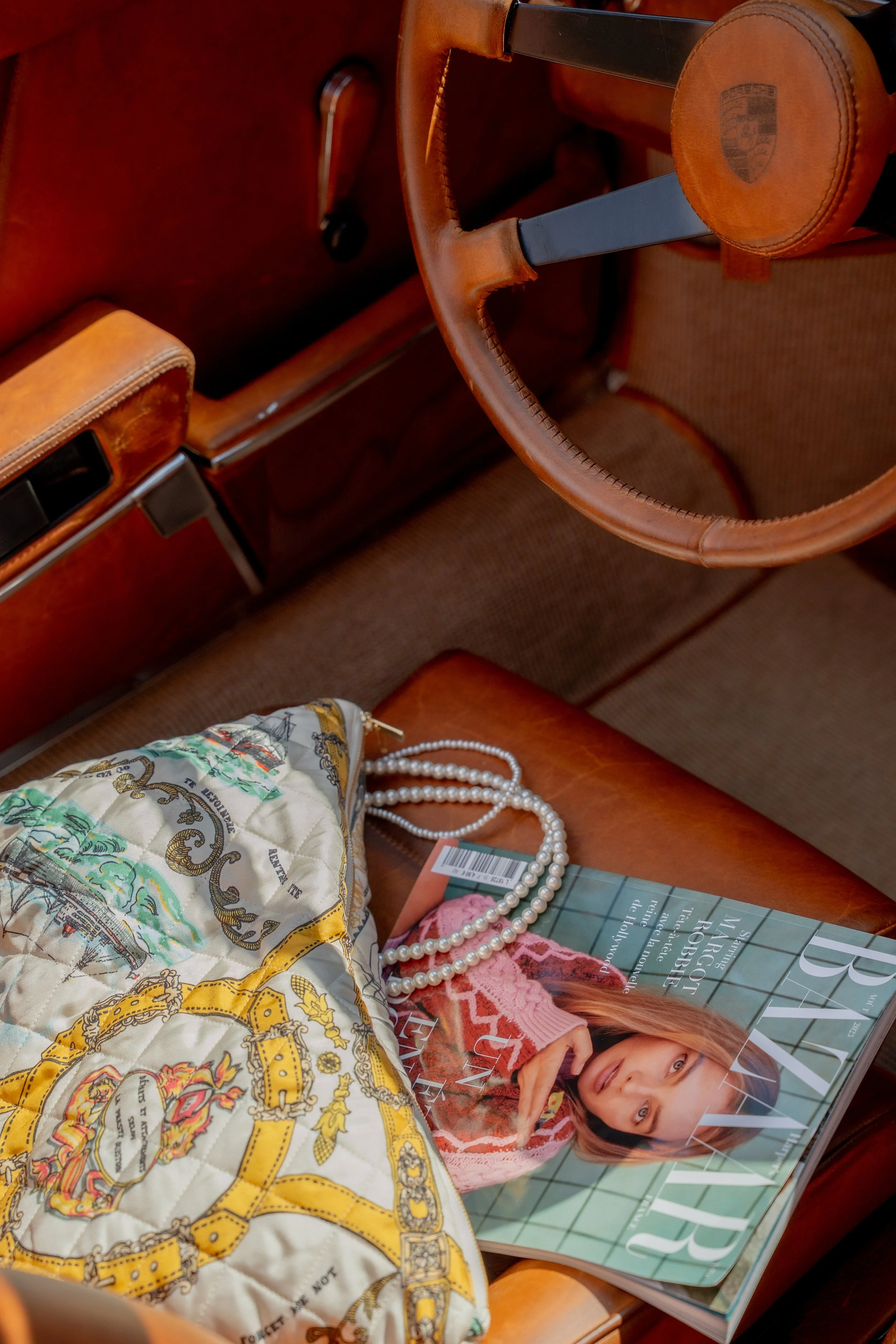Intérieur d'une voiture vintage en cuir marron, vue du siège avec un livre, un collier de perles et un sac de mode posé dessus.