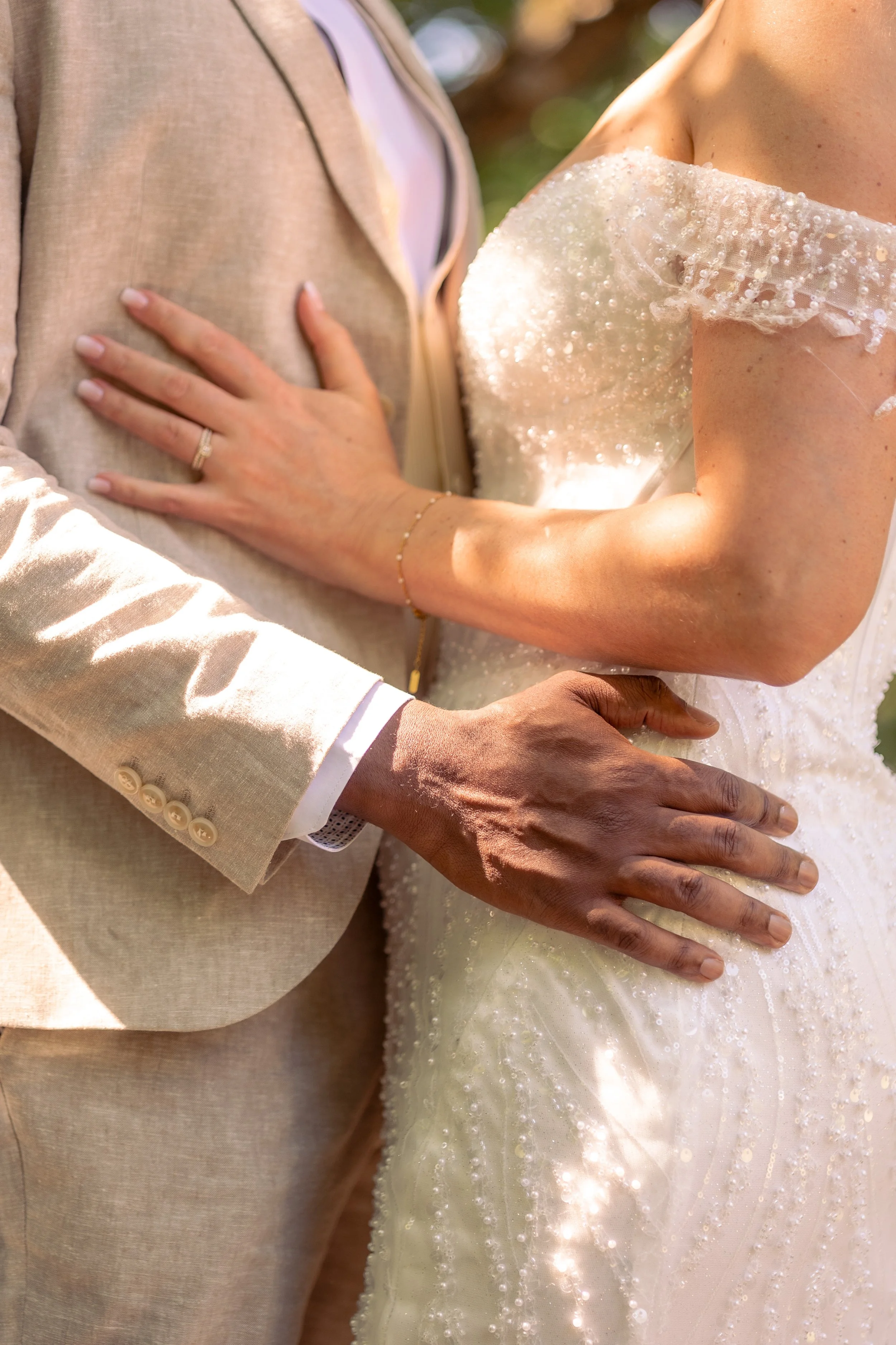 Gros plan sur un couple de mariés, le homme en costume beige clair, la femme en robe de mariage blanche avec des détails perlés, leurs mains jointes, lors d'une cérémonie nuptiale en plein air.