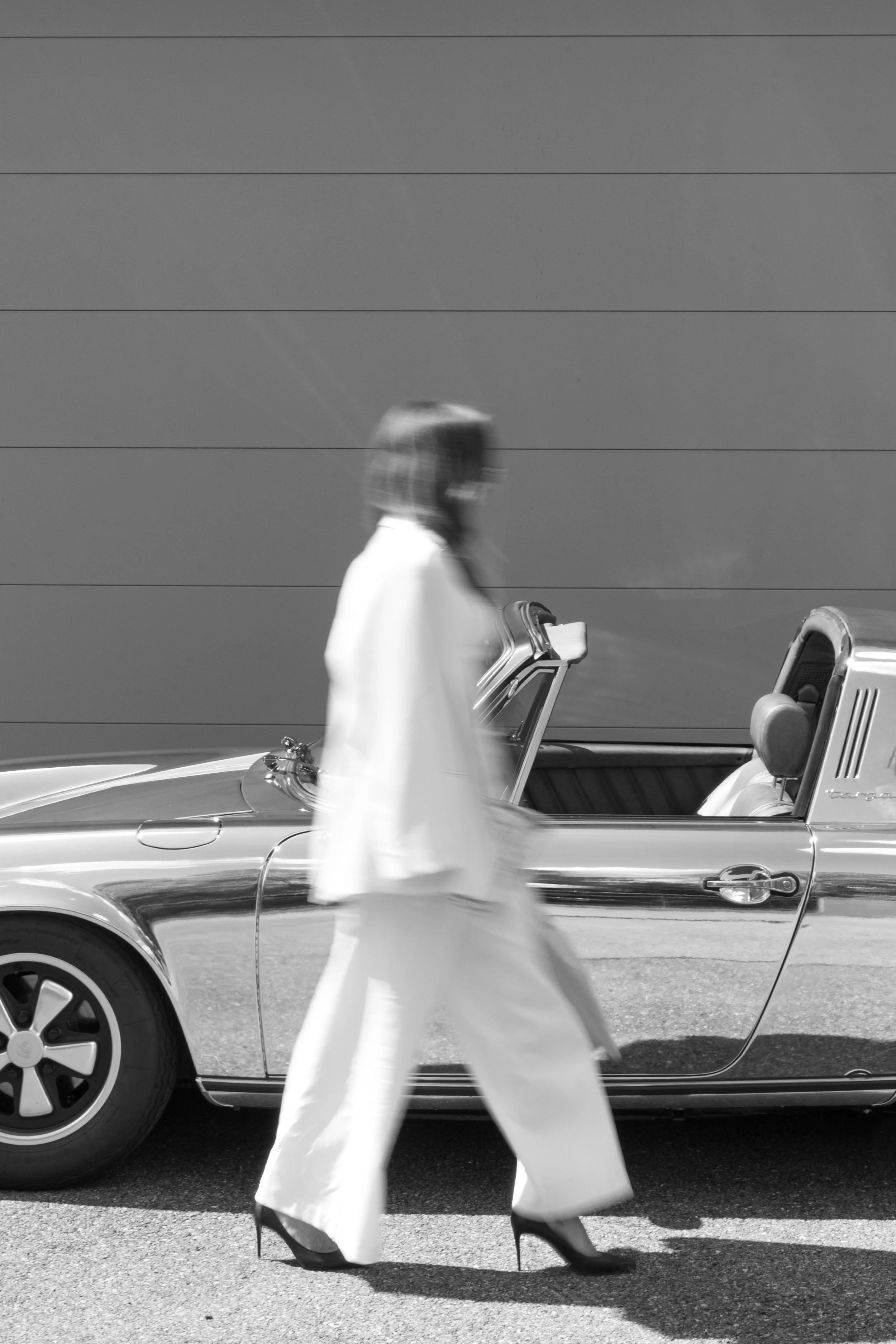 Une femme en vêtements blancs et talons hauts marche devant une voiture de sport décapotable stationnée devant un mur gris.